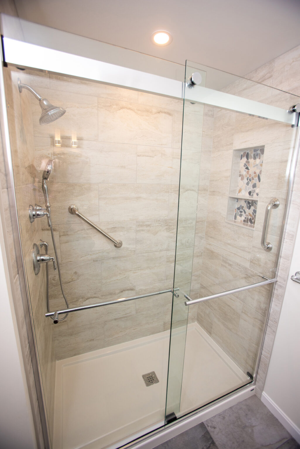 A modern shower with beige tile walls, a glass sliding door, overhead showerhead, hand-held shower, grab bars, and a decorative tile inset.