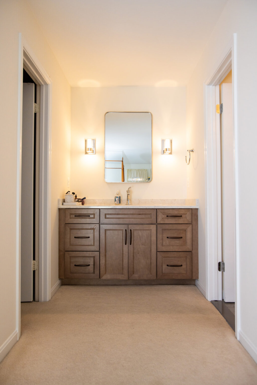 A bathroom with a wood vanity featuring a sink, a rectangular mirror, and two wall-mounted lights on either side. The area has cream-colored walls and doors on both sides.
