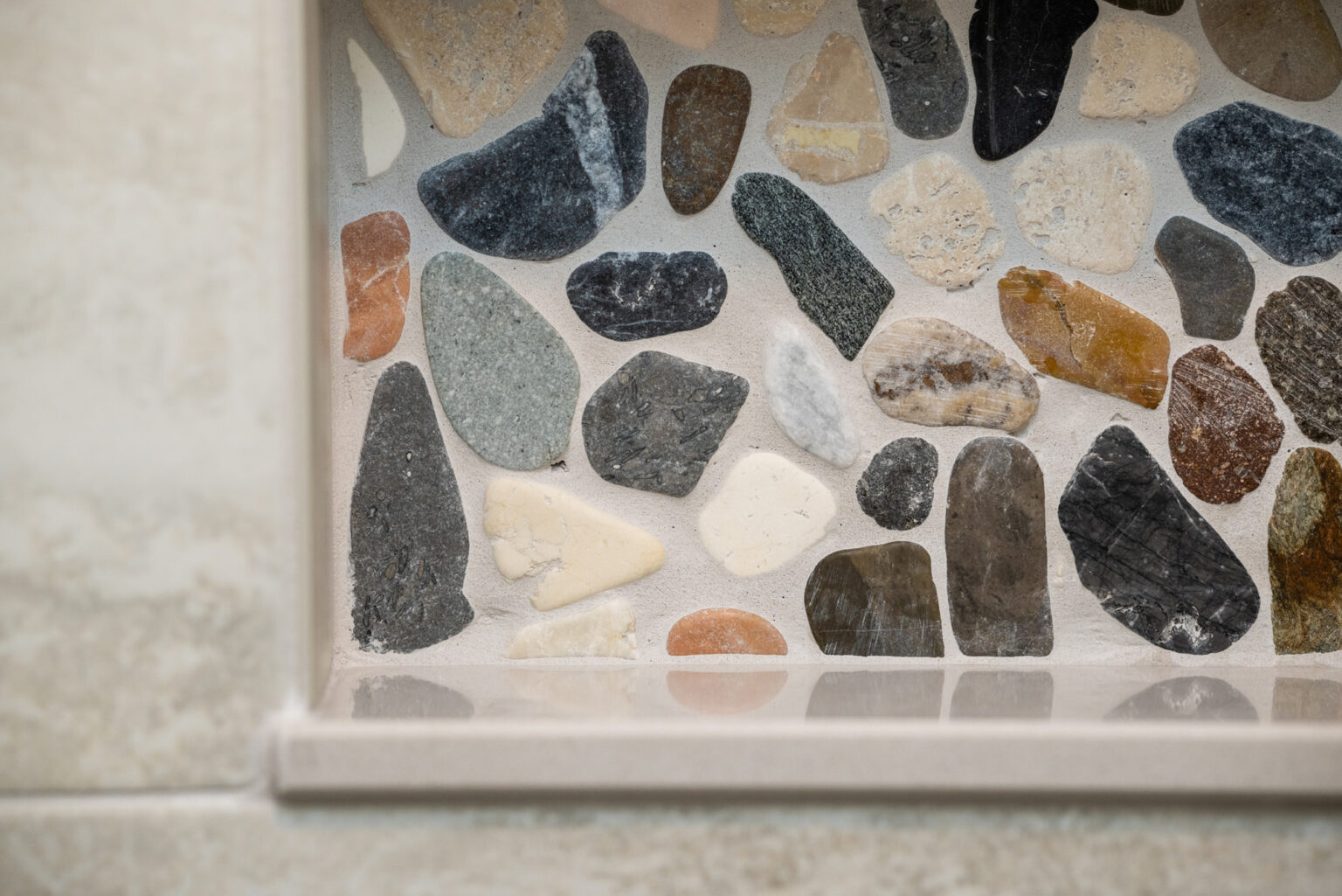 A close-up view of a mosaic made of various shapes and colors of smooth stones embedded in a rectangular recess, framed with light beige material.