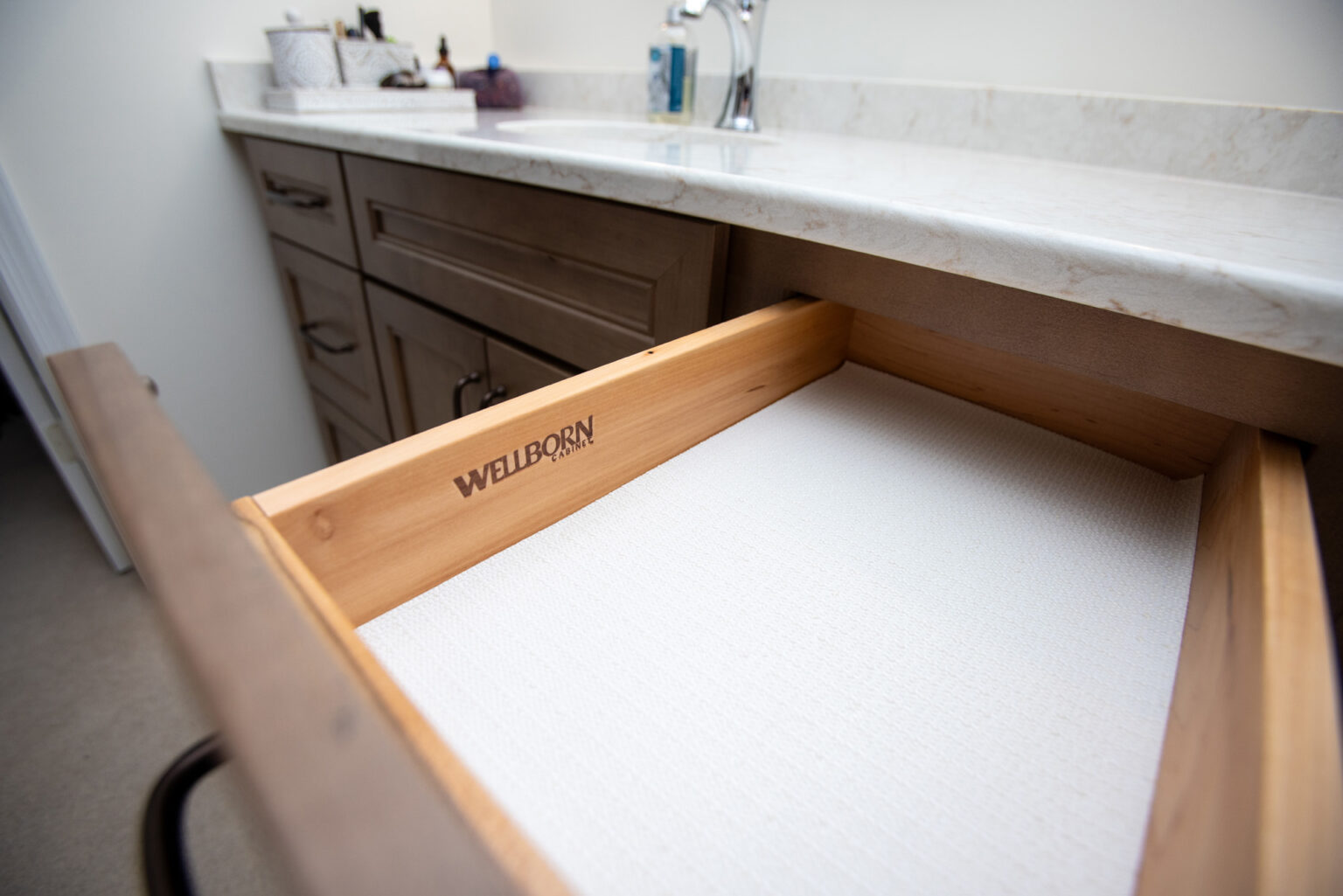 An open wooden drawer under a bathroom countertop labeled "Wellborn Cabinet" with a textured white lining.