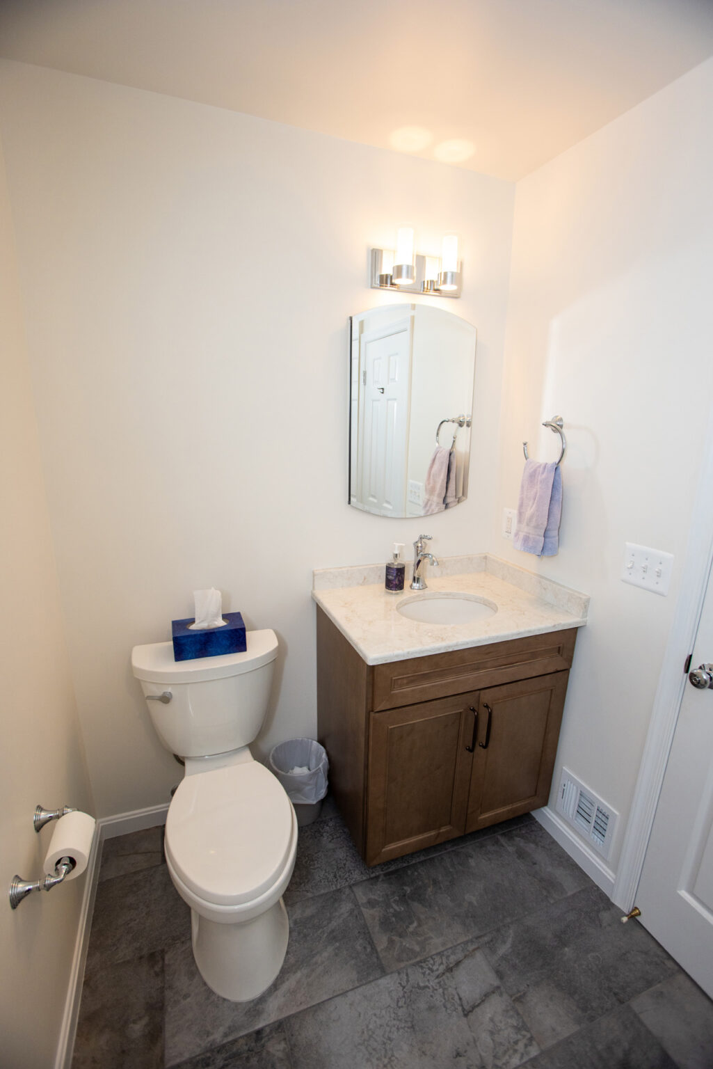A small bathroom with a toilet, a wooden vanity with a white countertop, a mirror, a towel ring with a purple towel, and tile flooring.