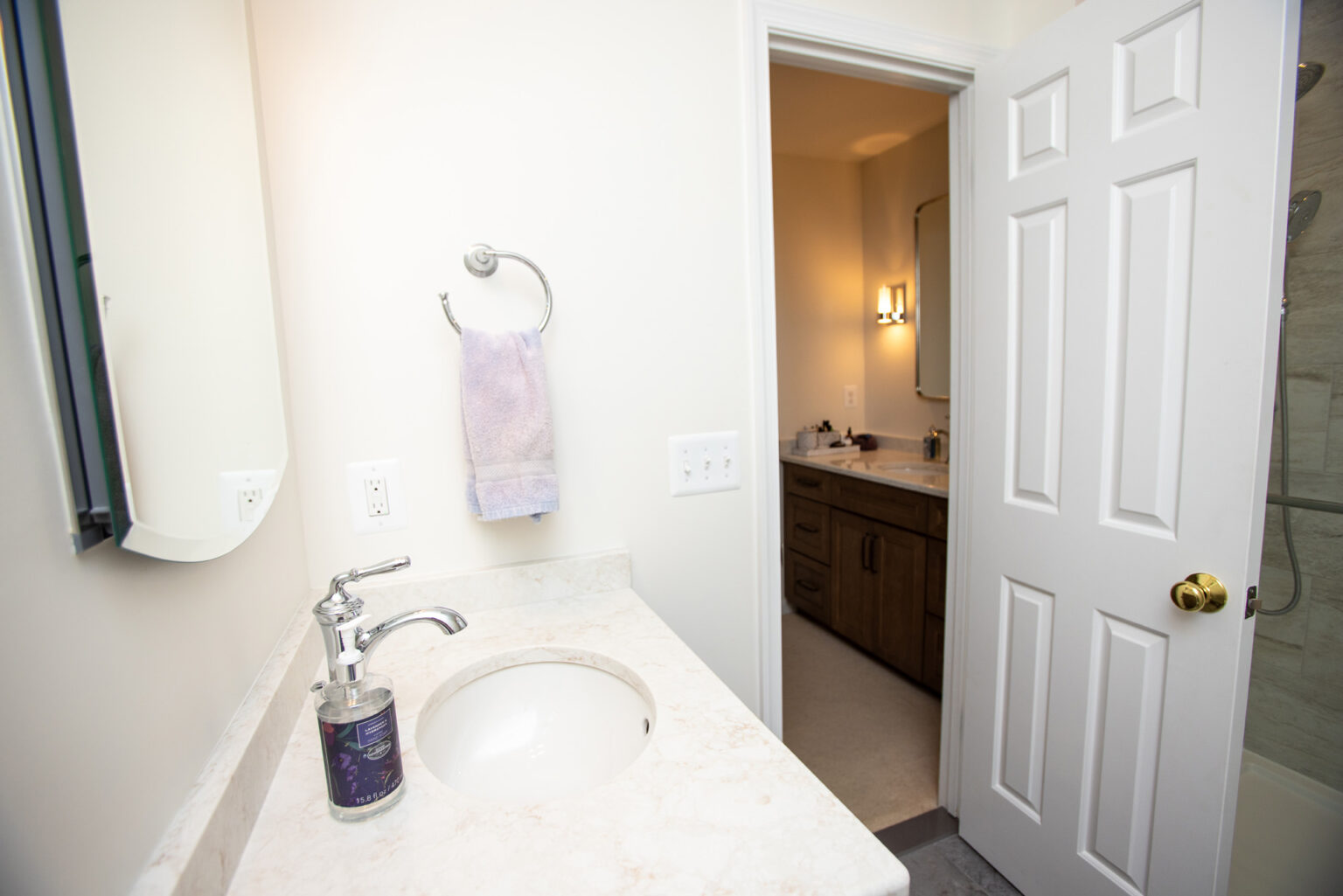 A bathroom with a white countertop, sink, soap dispenser, and towel on a ring. An open door leads to another part of the house with another bathroom area visible.