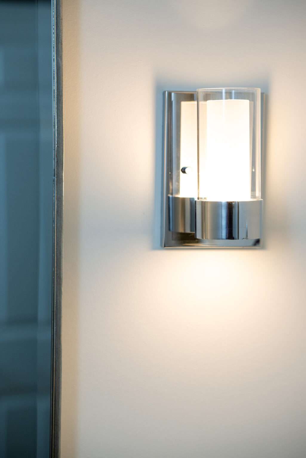 A modern wall-mounted light fixture with a metallic base and cylindrical glass cover, emitting a warm glow, is attached to a light-colored wall next to a mirror.
