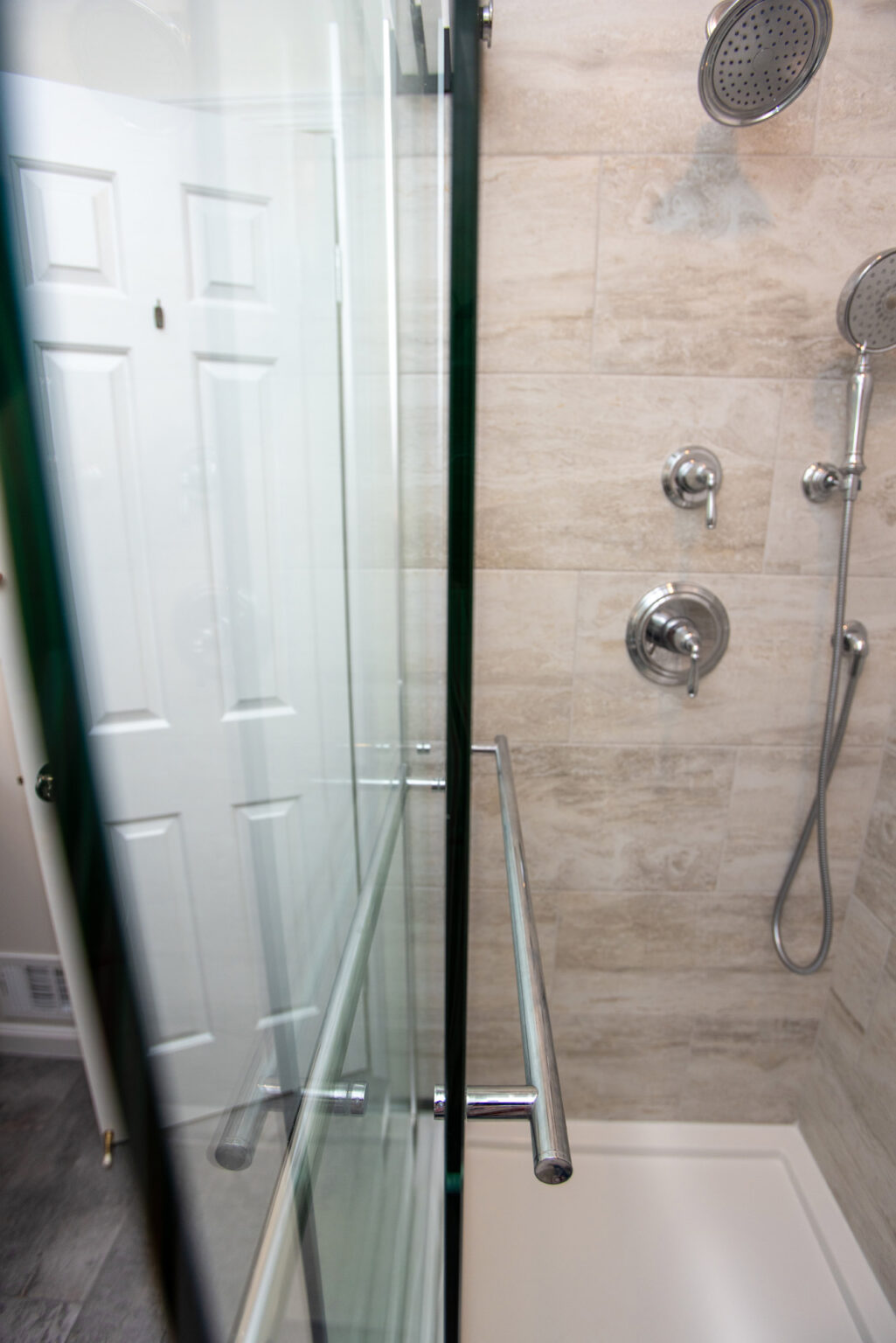 A modern shower with a glass door, chrome fixtures, a rain shower head, and a handheld shower head attached to a beige-tiled wall.