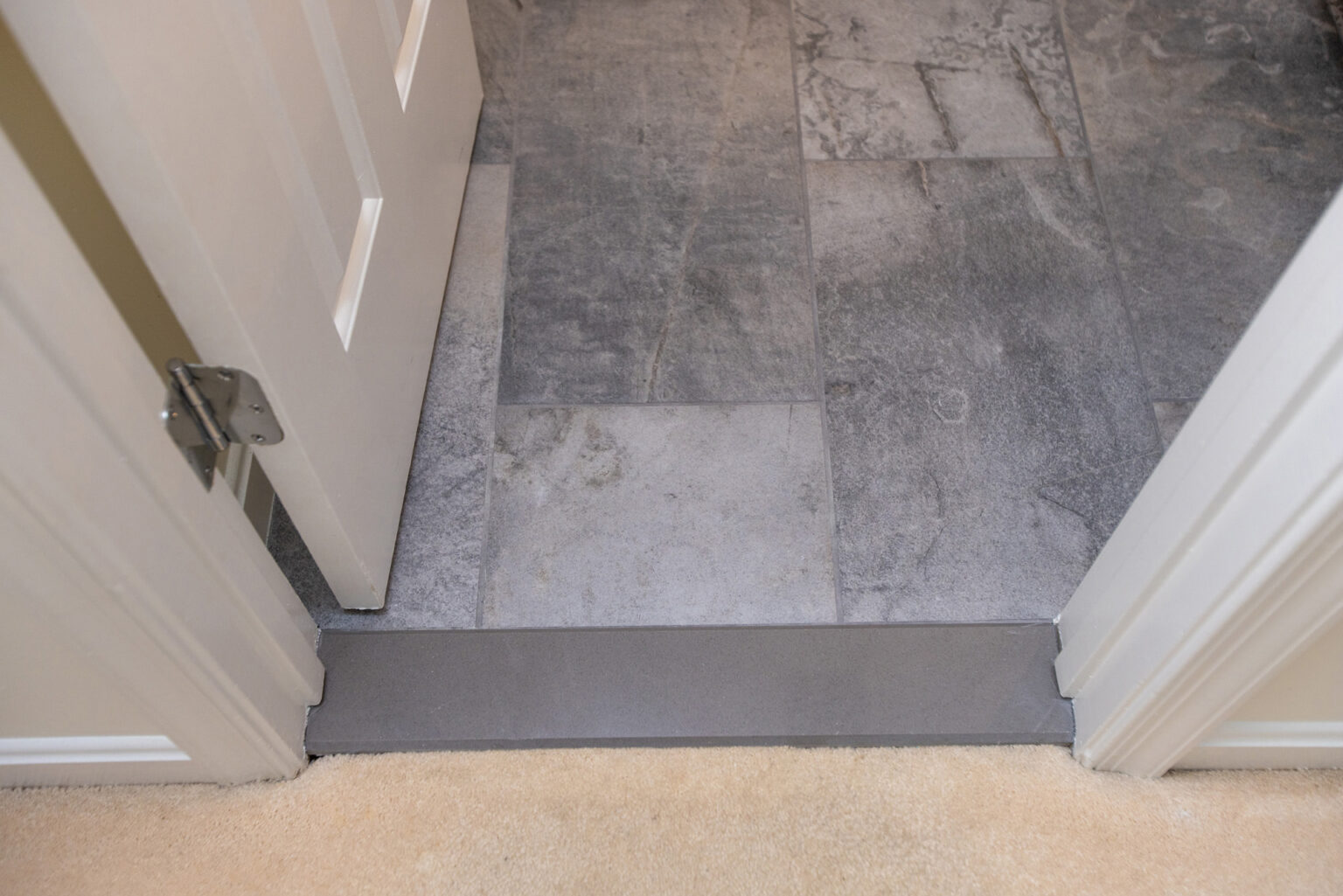 A doorway with gray tile flooring leading to a room with beige carpet. The door is partially open, revealing the contrasting flooring materials.