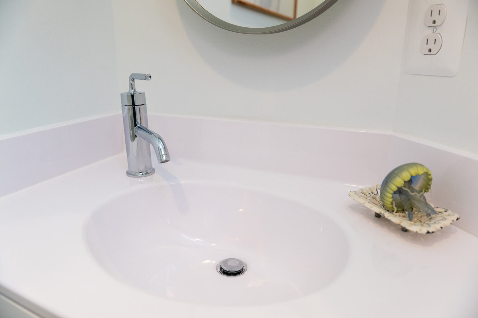 Image by Lesnick Photo A modern bathroom sink with a silver faucet, a round mirror above, and a soap dish containing a green dental retainer on the counter.