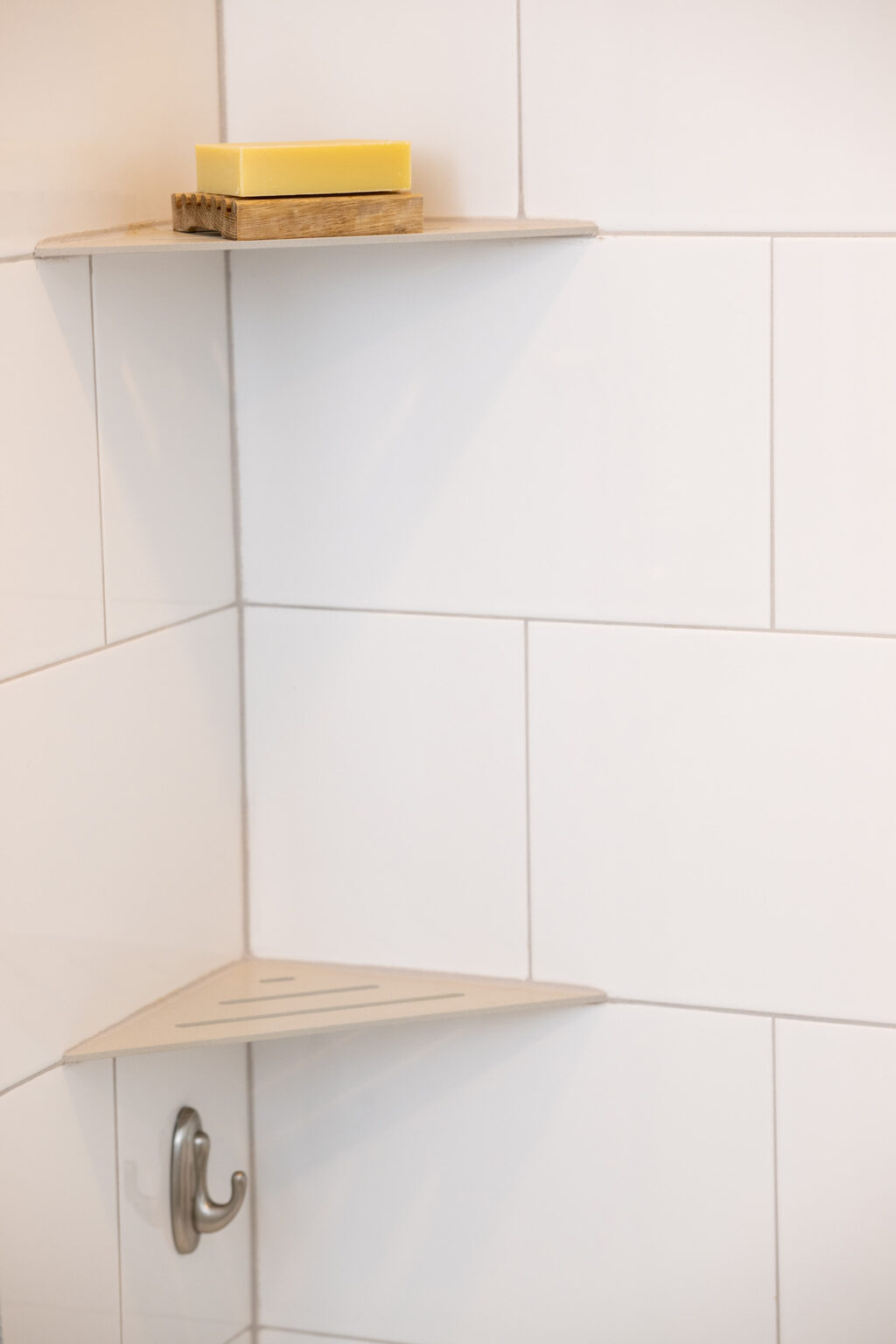 Image by Lesnick Photo A shower corner shelf with a wooden soap dish holding a bar of yellow soap. A metal hook is attached below the shelf to the white tiled wall.