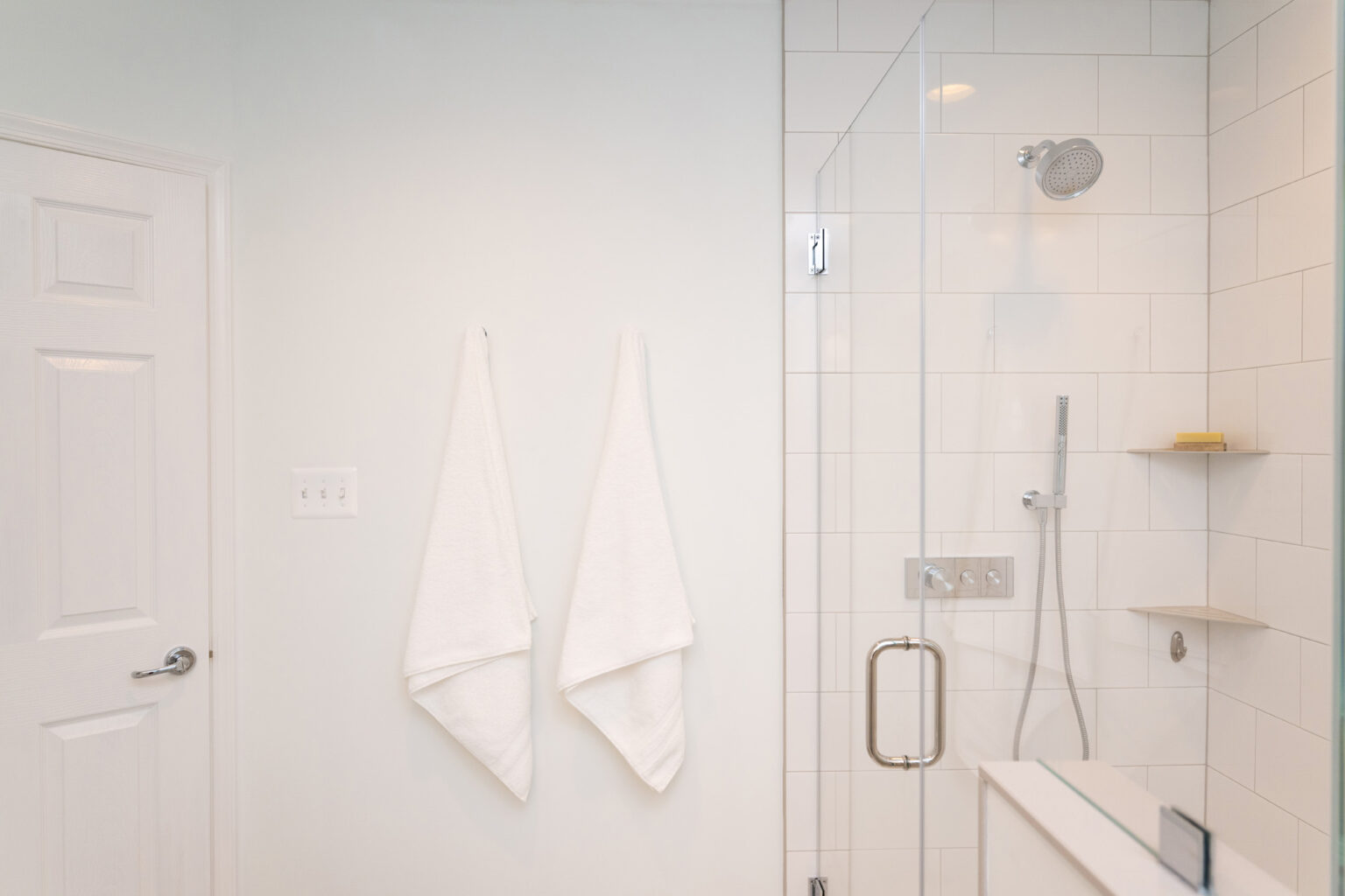 Image by Lesnick Photo A modern bathroom featuring a glass-enclosed shower, white tiled walls, two white towels hanging on hooks, and a closed white door.