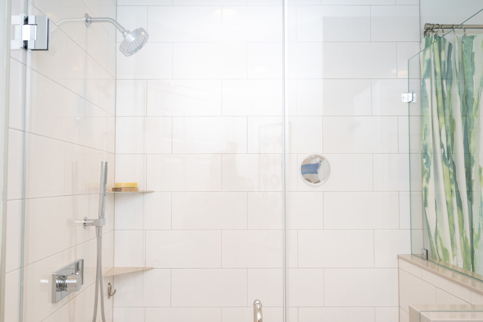 Image by Lesnick Photo A modern white-tiled shower with a glass door, wall-mounted showerhead, and a handheld showerhead. A small shelf holds soap, and a green-striped shower curtain is partially visible on the right.