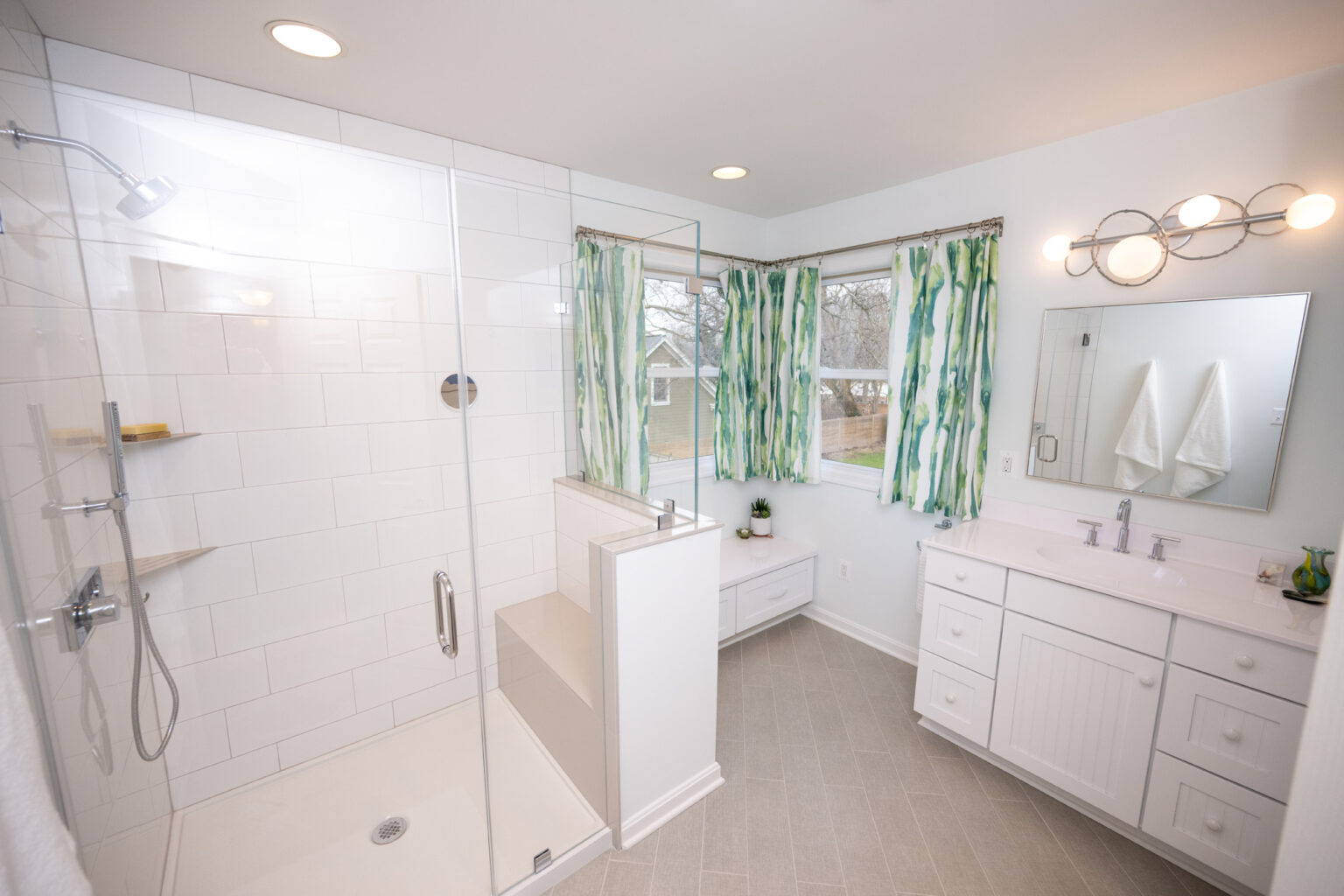 Image by Lesnick Photo A bathroom with a glass-enclosed shower, green and white curtains, a large mirror above a white vanity, and a seating area under the window.