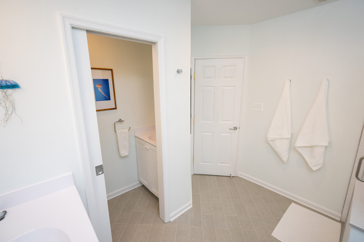 Image by Lesnick Photo A clean, white bathroom featuring a vanity with small sink, two hanging white towels, a door, and a framed picture on the wall.
