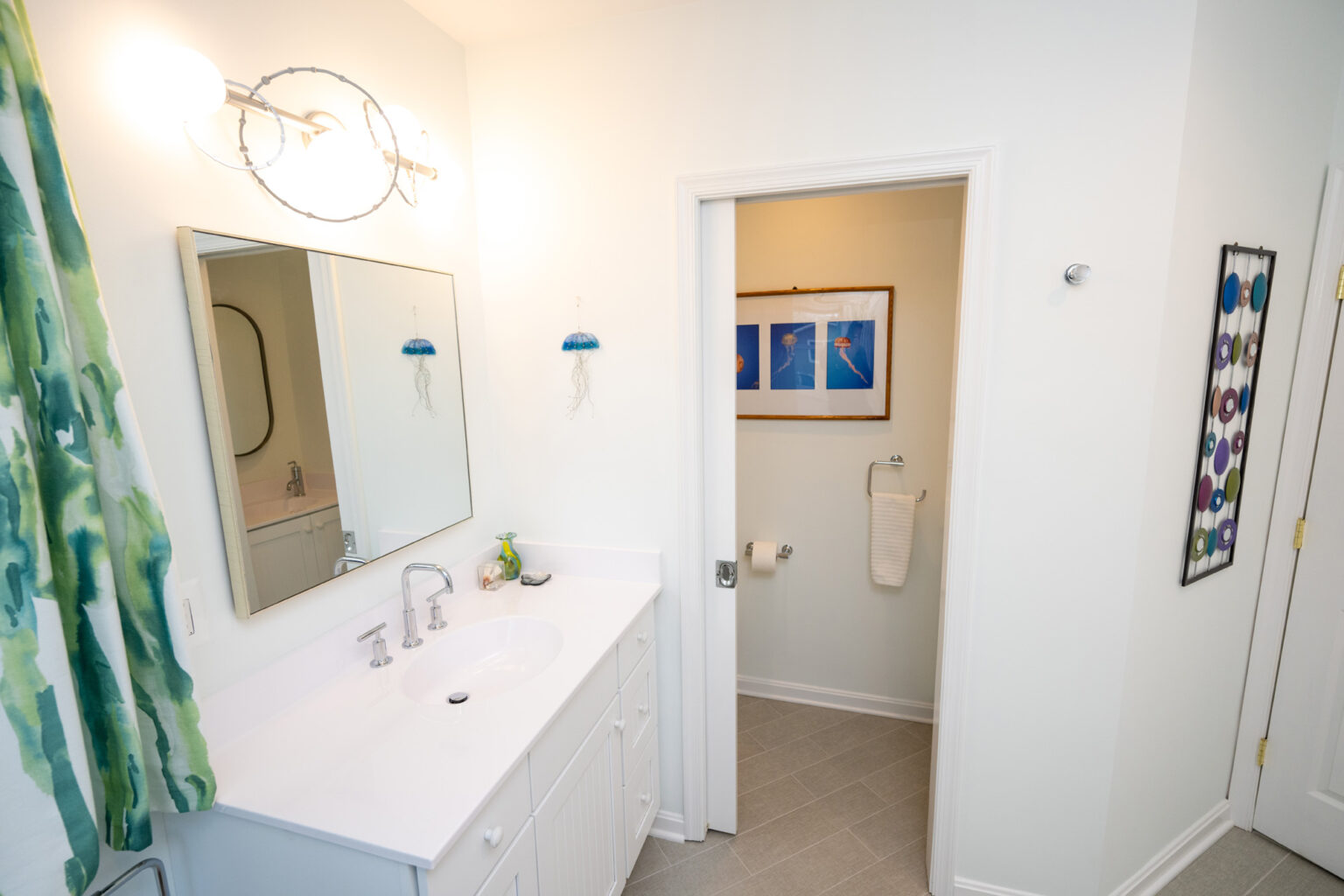 Image by Lesnick Photo A bathroom with a sink, vanity mirror, and wall-mounted light on the left. A door leads to a toilet area with framed pictures on the wall and a towel hanging on a ring. A patterned shower curtain is visible.