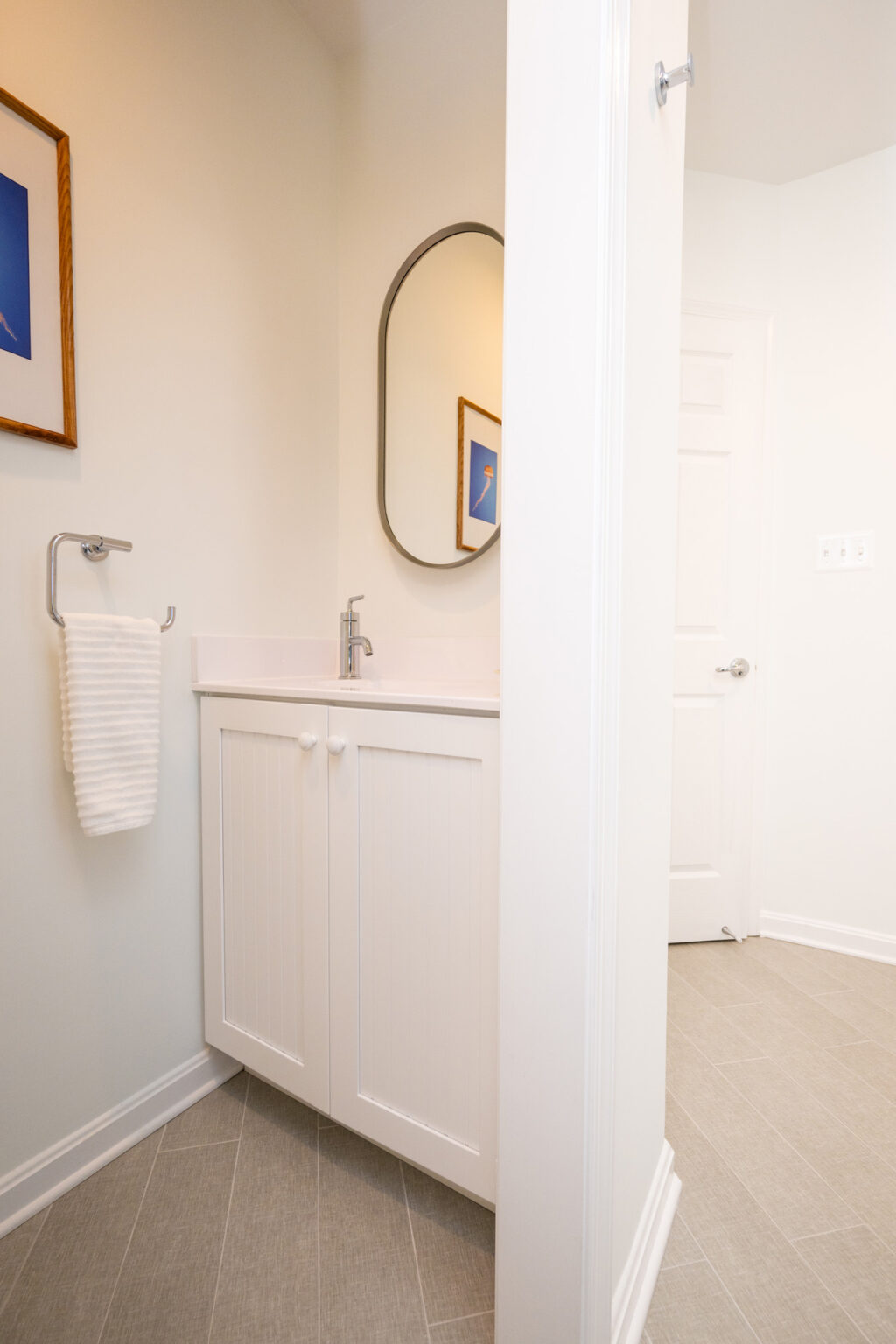 Image by Lesnick Photo A small bathroom with a white vanity and sink, a circular mirror, a hand towel on a holder, and framed artwork on the wall. The floor is tiled, and a door is partially visible to the right.