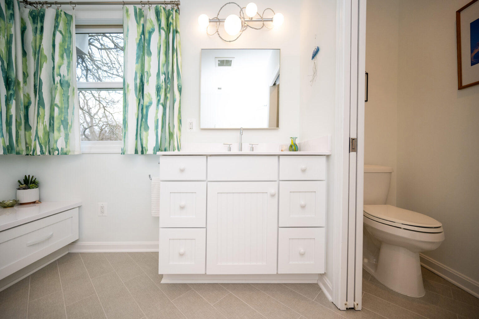 Image by Lesnick Photo A clean, modern bathroom with a white vanity and sink, green patterned curtains, a large mirror, and a toilet in the corner. The floor is tiled in light gray.