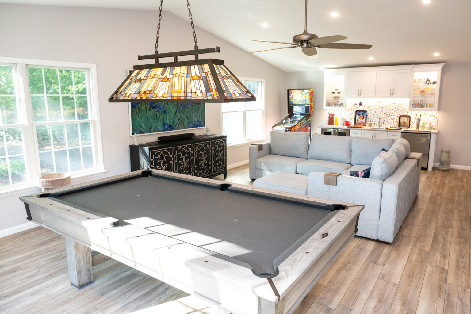 Image by Lesnick Photo Modern entertainment room with a pool table, L-shaped sofa, pinball machine, and bar area. There are large windows, a ceiling fan, and overhead lighting, including a stained glass fixture above the pool table.
