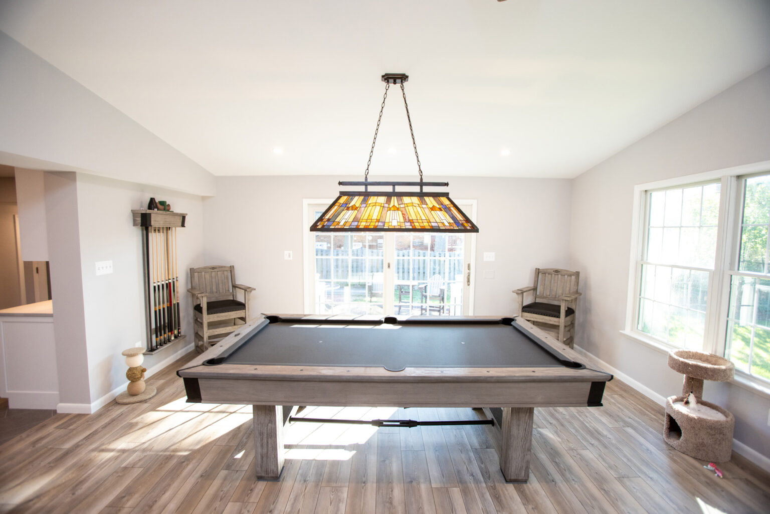 Image by Lesnick Photo A well-lit game room features a pool table under a large stained-glass light fixture. The room includes hardwood floors, tall windows, and a pool cue rack with cues in the corner.