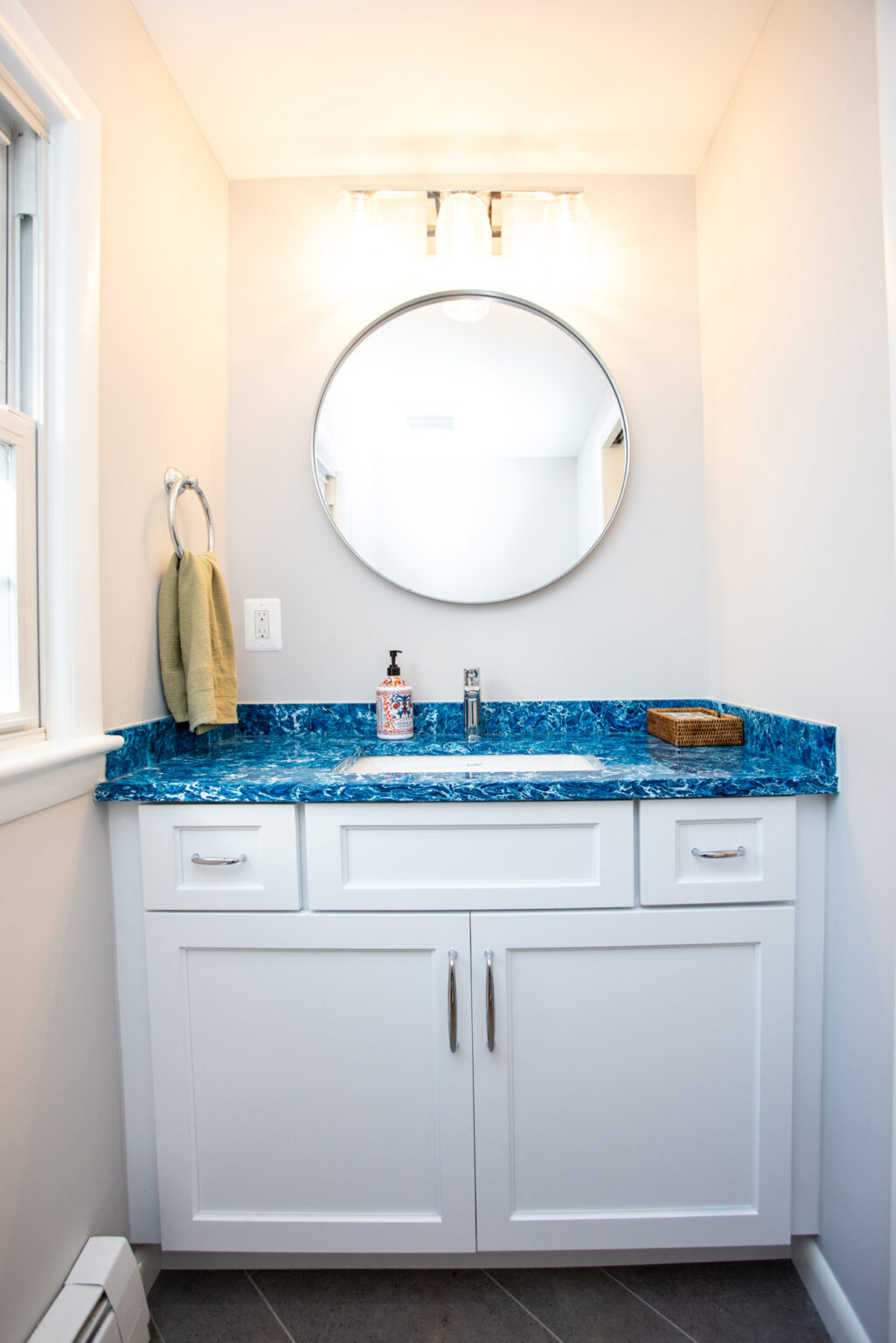 Image by Lesnick Photo A small bathroom vanity with white cabinets, a blue countertop, a circular mirror, a soap dispenser, a wicker basket, and a hand towel on a ring holder.