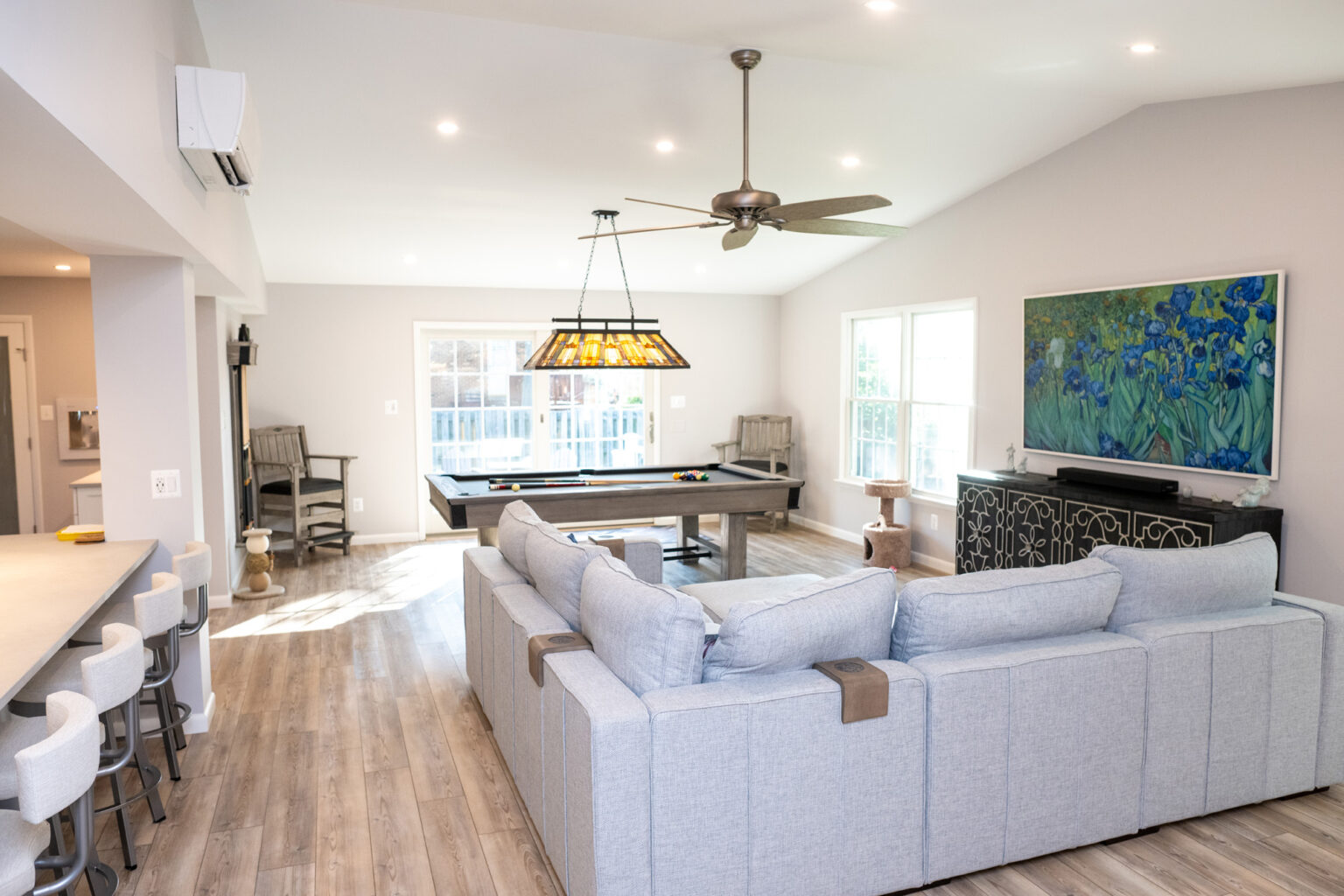 Image by Lesnick Photo A spacious living room with a gray sectional couch, a pool table, a ceiling fan, and a large painting on the wall. The room has wooden flooring and large windows allowing natural light to enter.