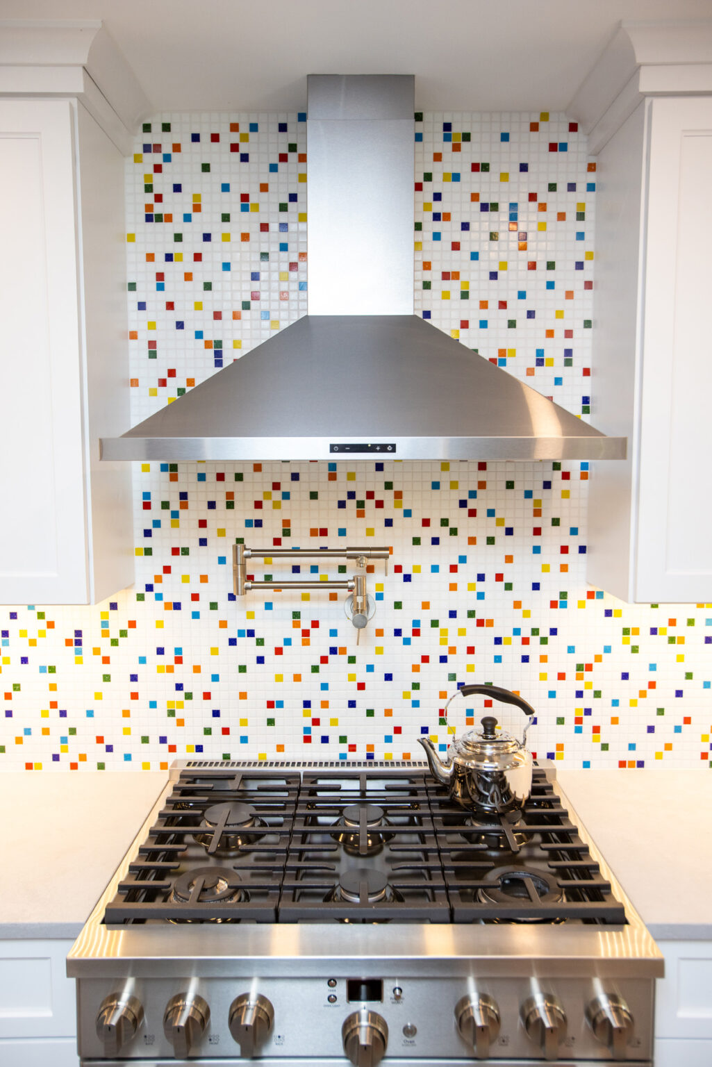 Image by Lesnick Photo A stainless steel range hood above a stove with a kettle. The wall features a colorful mosaic tile backsplash with white cabinets on either side.