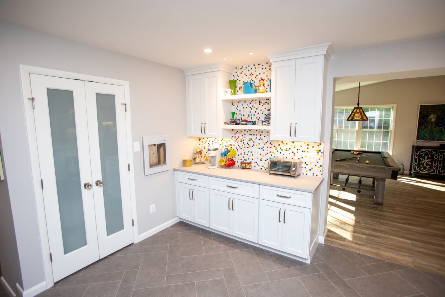 Image by Lesnick Photo A modern kitchen corner with white cabinets, colorful backsplash, floating shelves, and recessed lighting. To the right is a doorway opening to a room with a pool table and hanging light fixture.