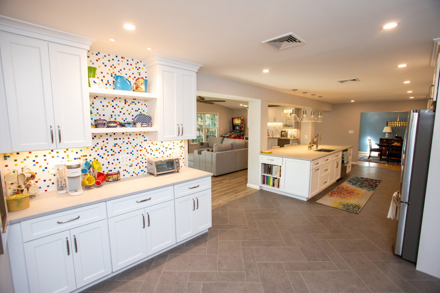 Image by Lesnick Photo Bright modern kitchen with white cabinetry, colorful mosaic backsplash, and spacious layout leading to a living area. A large central island has a sink and hanging pendant lights above.