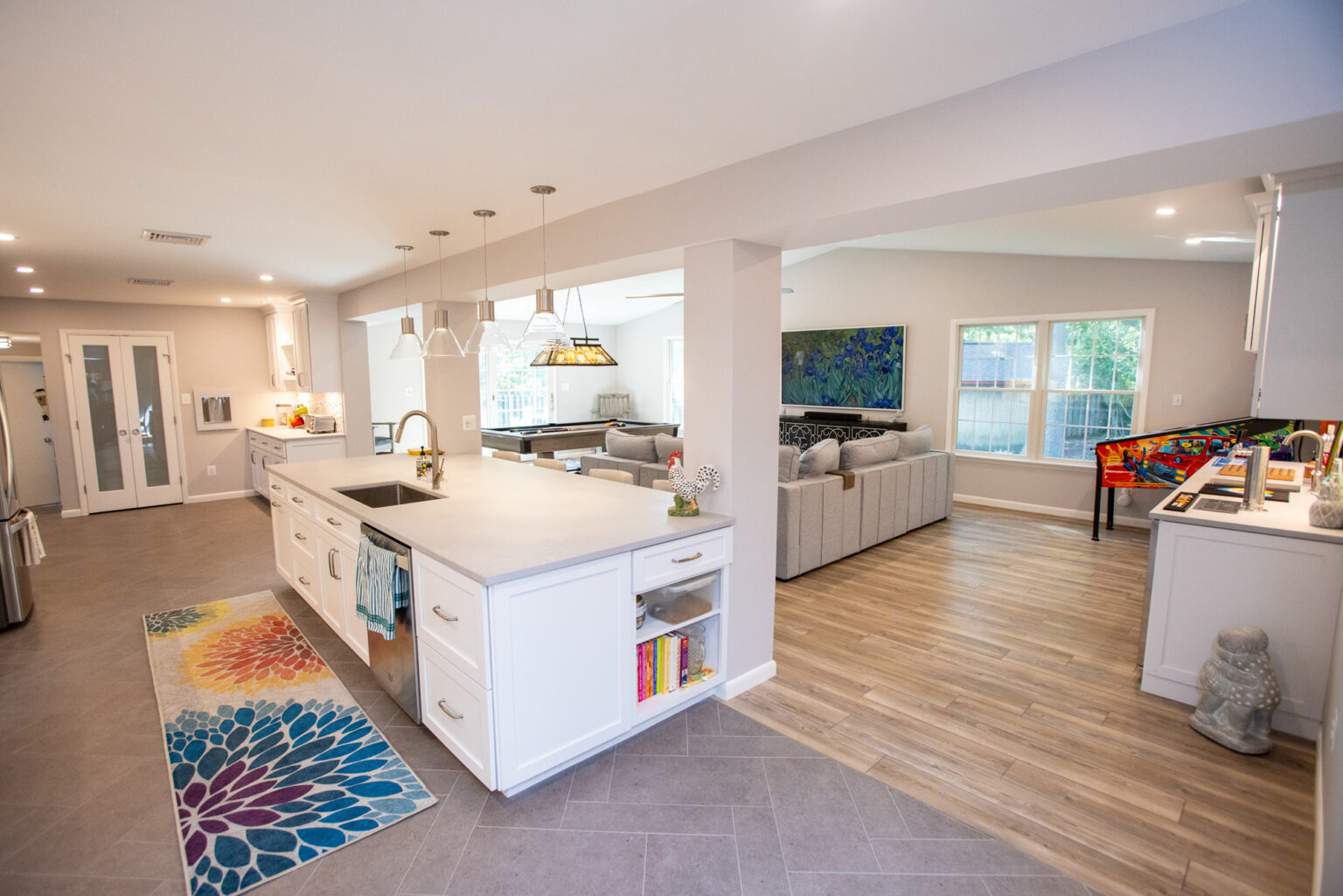 Image by Lesnick Photo A modern open-concept living space featuring a kitchen island with pendant lights, a colorful area rug, a large sectional sofa in the living area, and a pinball machine by the windows.