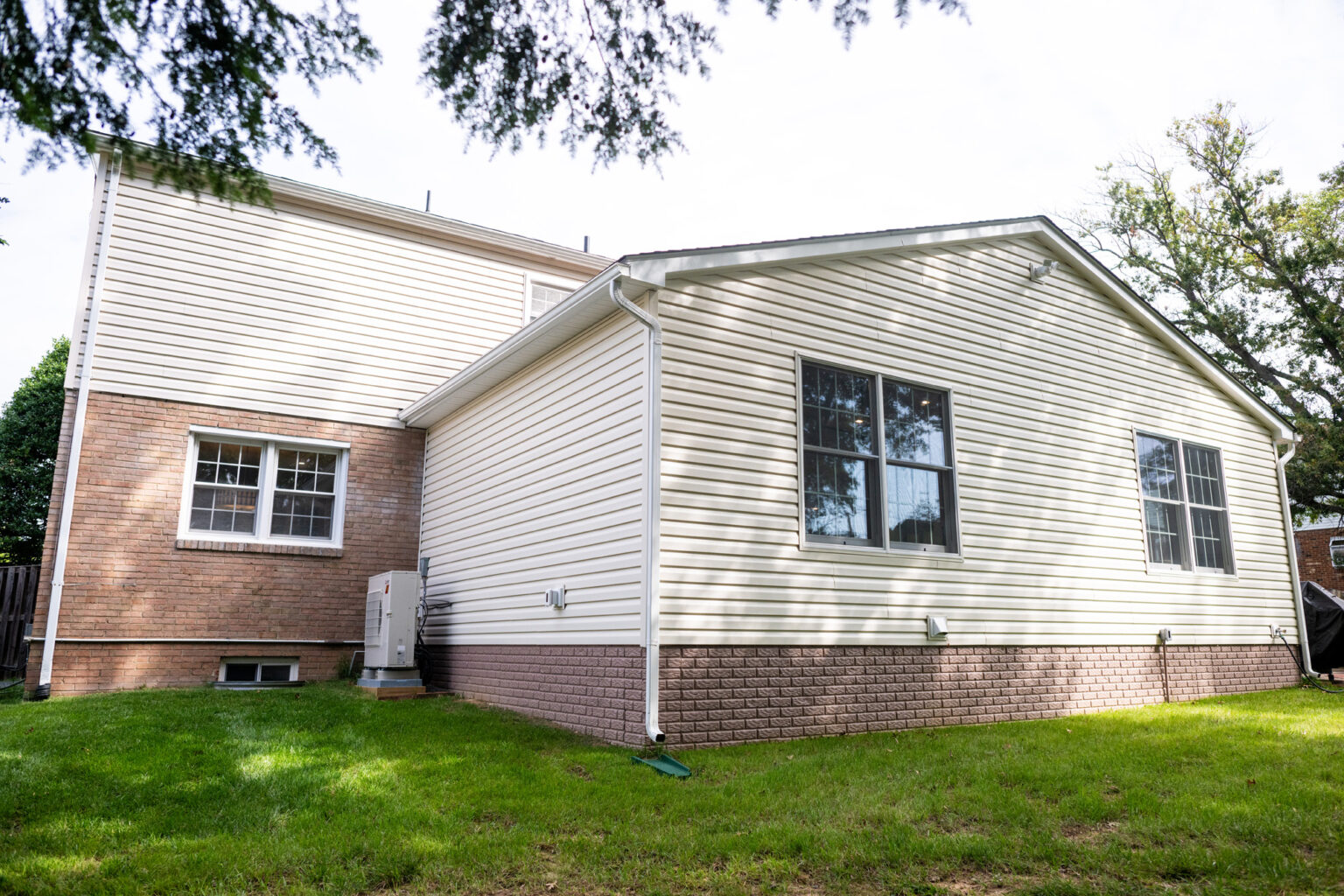 Image by Lesnick Photo A two-story house with beige siding and brick accents, featuring multiple windows and a well-maintained lawn under a partly cloudy sky.