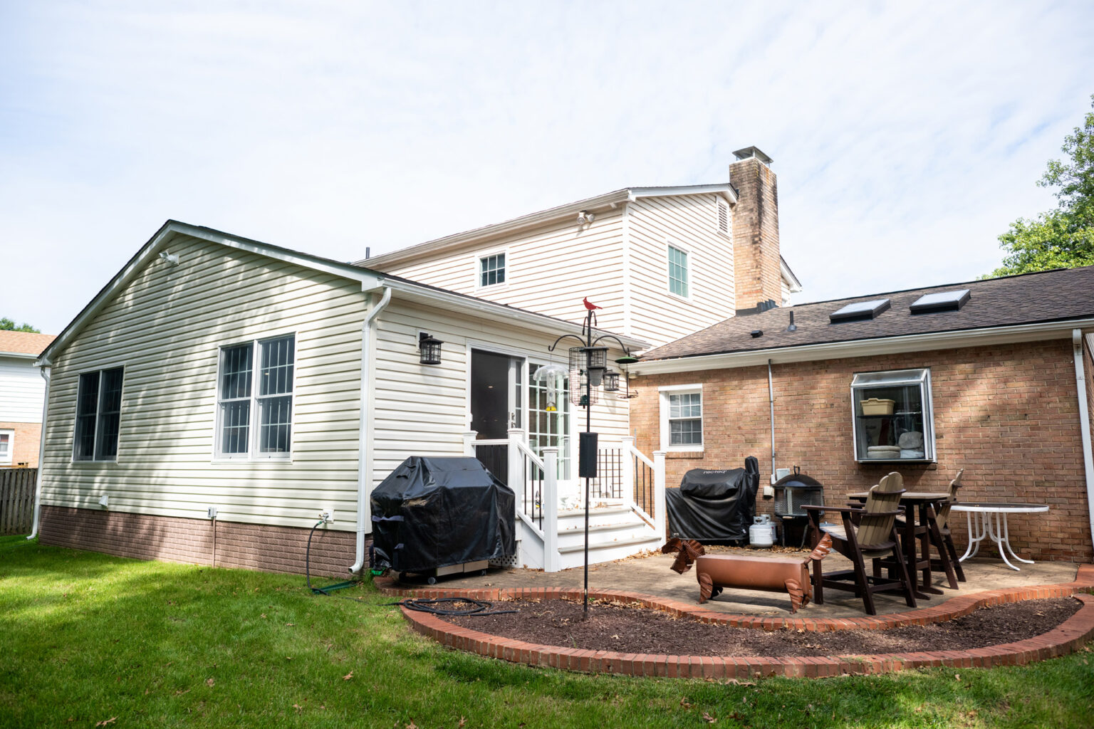 Image by Lesnick Photo A backyard with a back entrance to a house, a grill, a bird feeder, a patio with dining furniture, skylights, and a small area with potted plants.