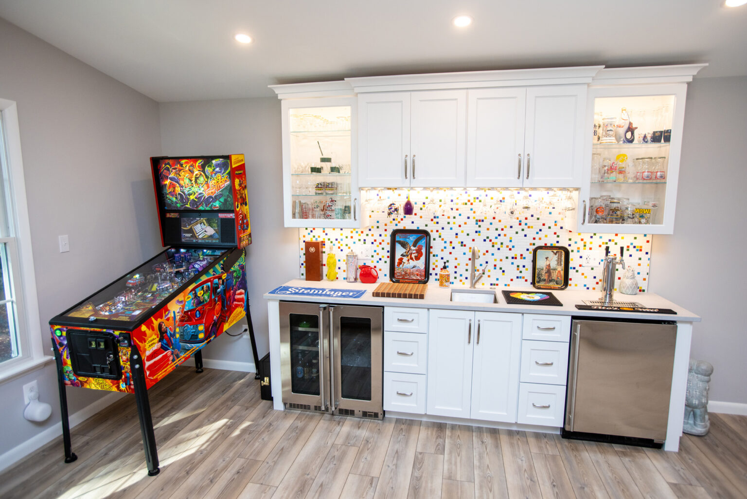Image by Lesnick Photo A modern game room with a pinball machine, a kitchenette featuring white cabinets, a colorful tiled backsplash, a dishwasher, and a mini fridge. Shelves display various items and glassware.