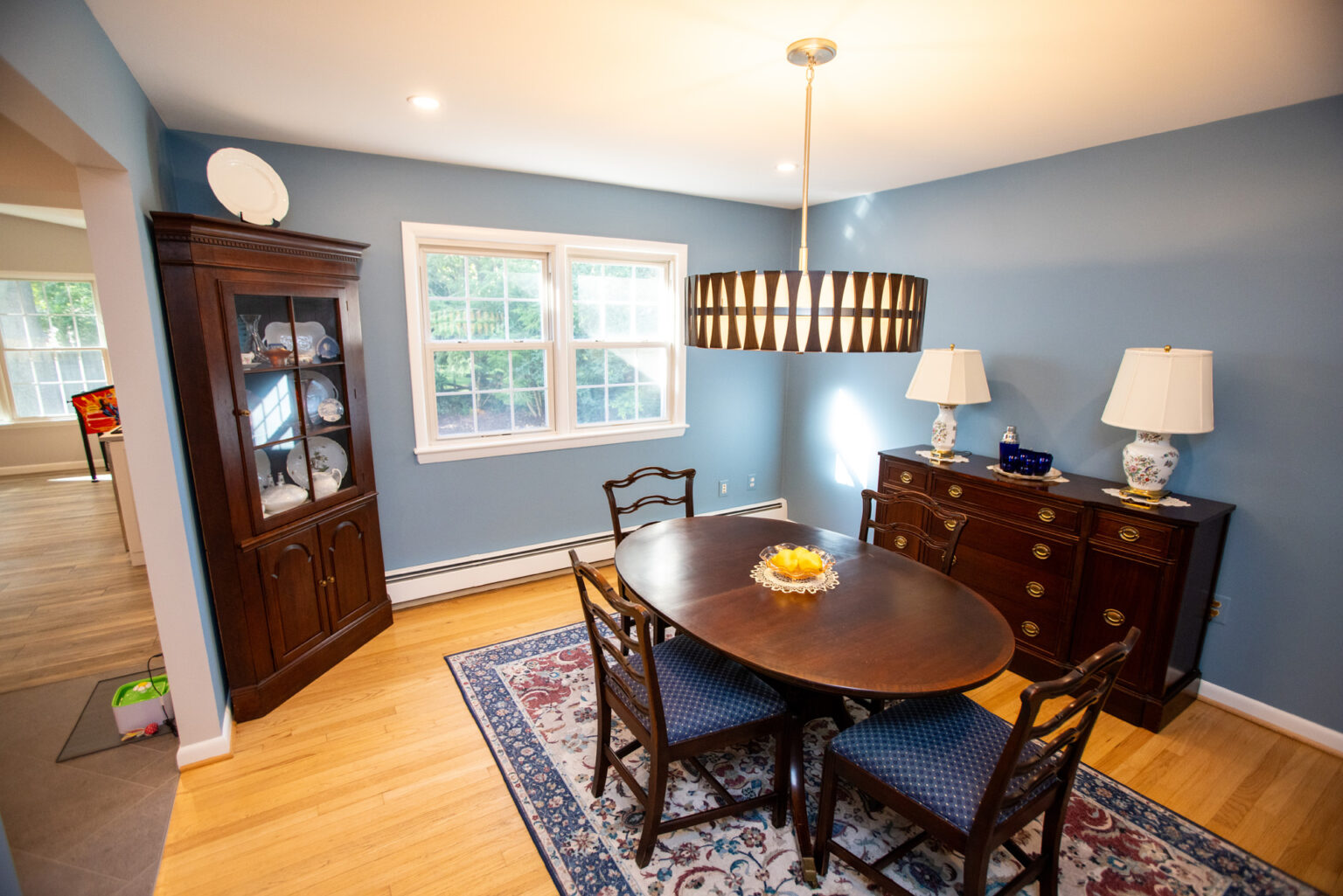 Image by Lesnick Photo A dining room with blue walls, a wooden table with four chairs, a patterned rug, a sideboard with two lamps, a display cabinet with dishes, and a large window. A chandelier hangs above the table.