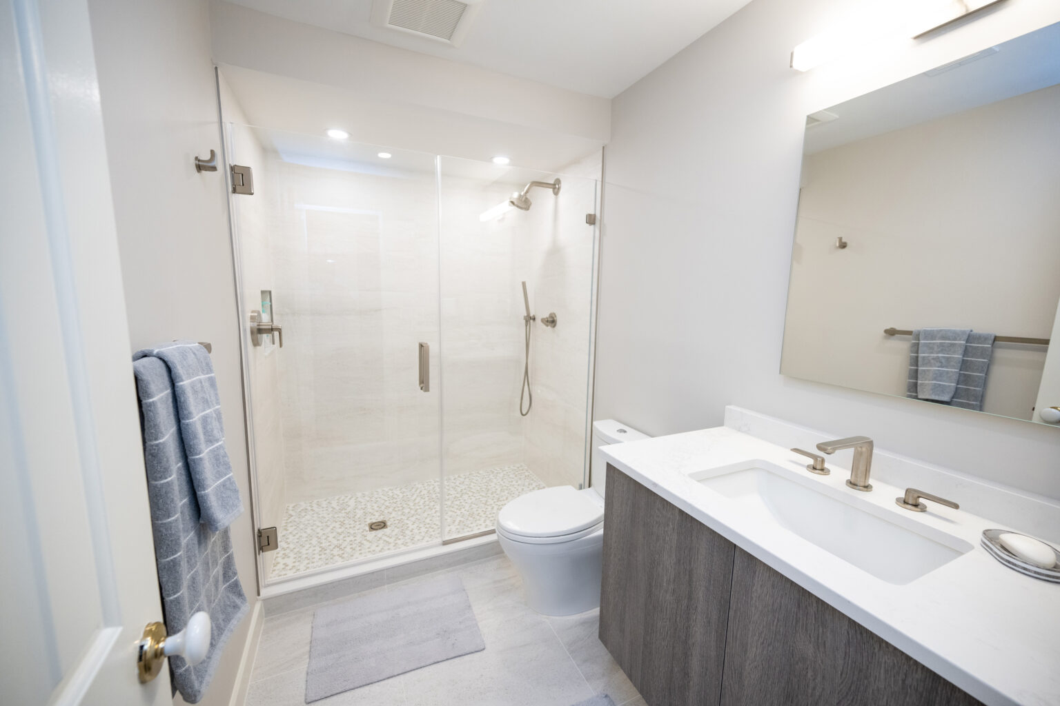 Modern bathroom with a glass-enclosed shower, white toilet, large mirror, sink with storage cabinet, towel rack holding folded towels, and overhead lighting. The floor is tiled in light gray.