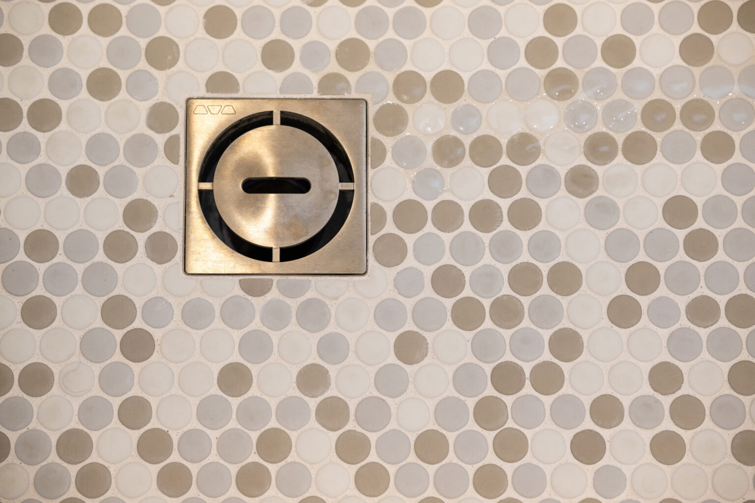 A metal drain cover is set in a wall with small round tiles in shades of white and light beige.
