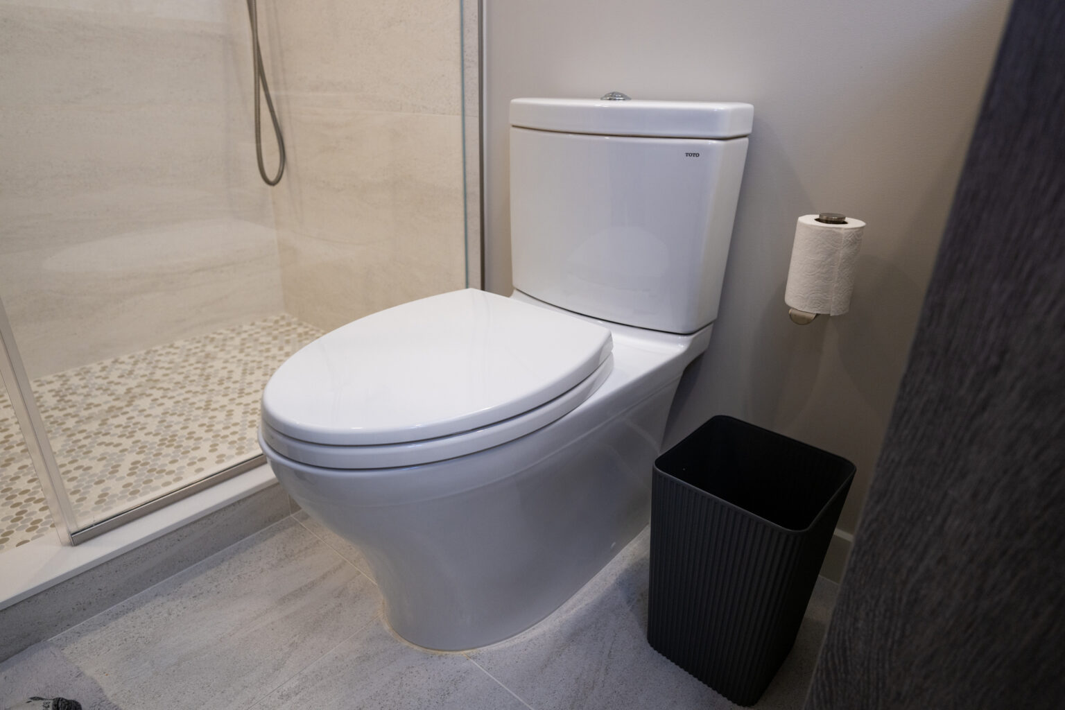 A modern bathroom featuring a white toilet, a roll of toilet paper on a holder, a black waste bin, and a shower with a glass wall and a tiled floor.