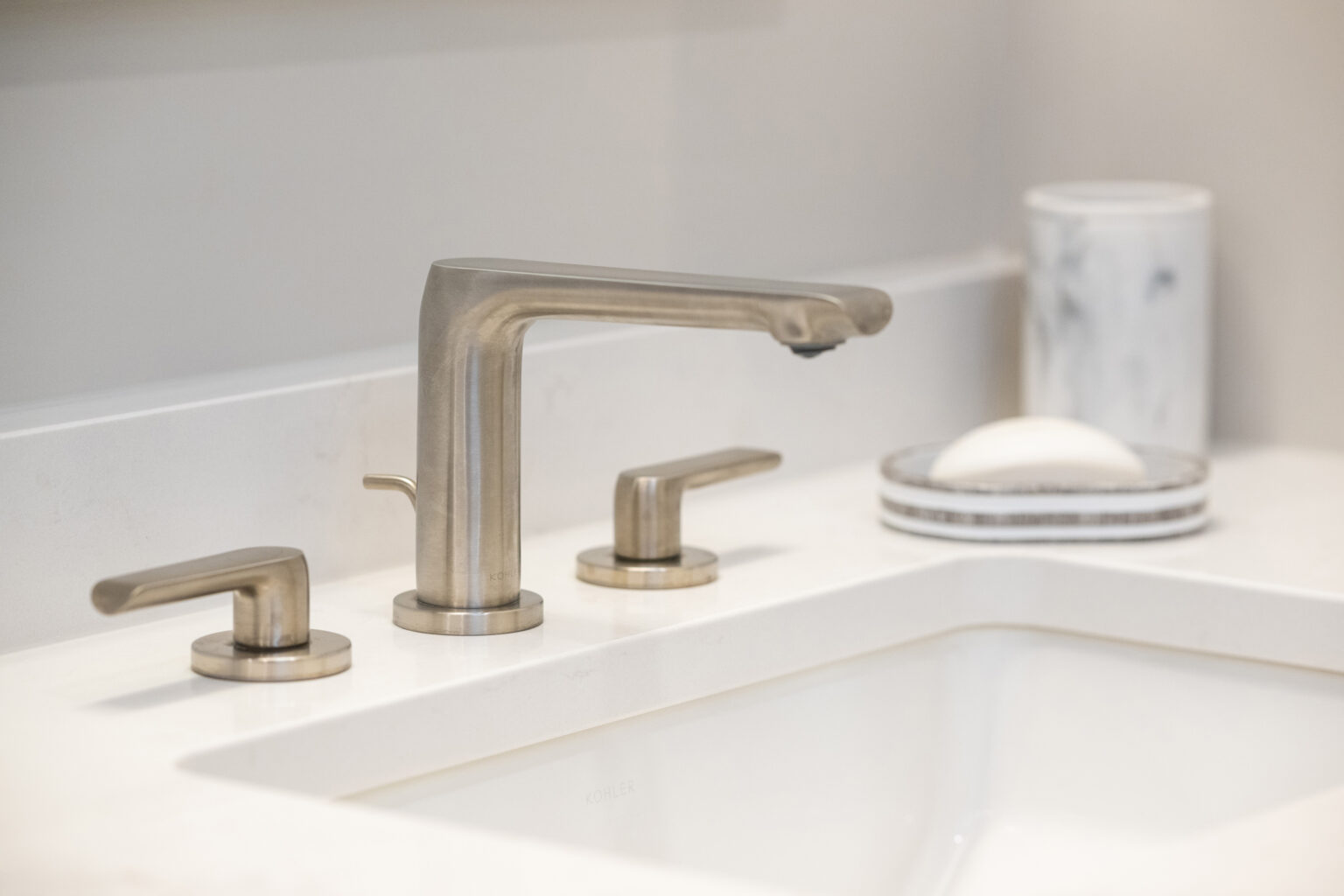 A close-up of a modern sink with a sleek faucet, two handles, a bar of soap in a marble dish, and a marble cup in the background. The sink is set in a white countertop.