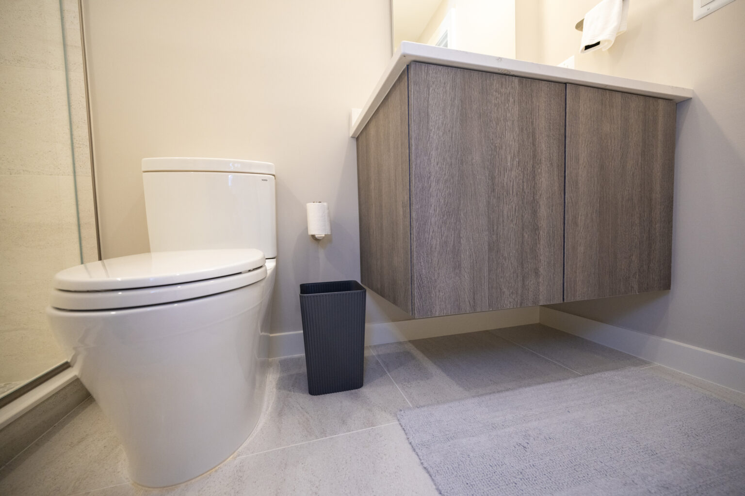 A modern bathroom with a white toilet, gray trash bin, floating wooden vanity, and a light-colored rug on the tiled floor.