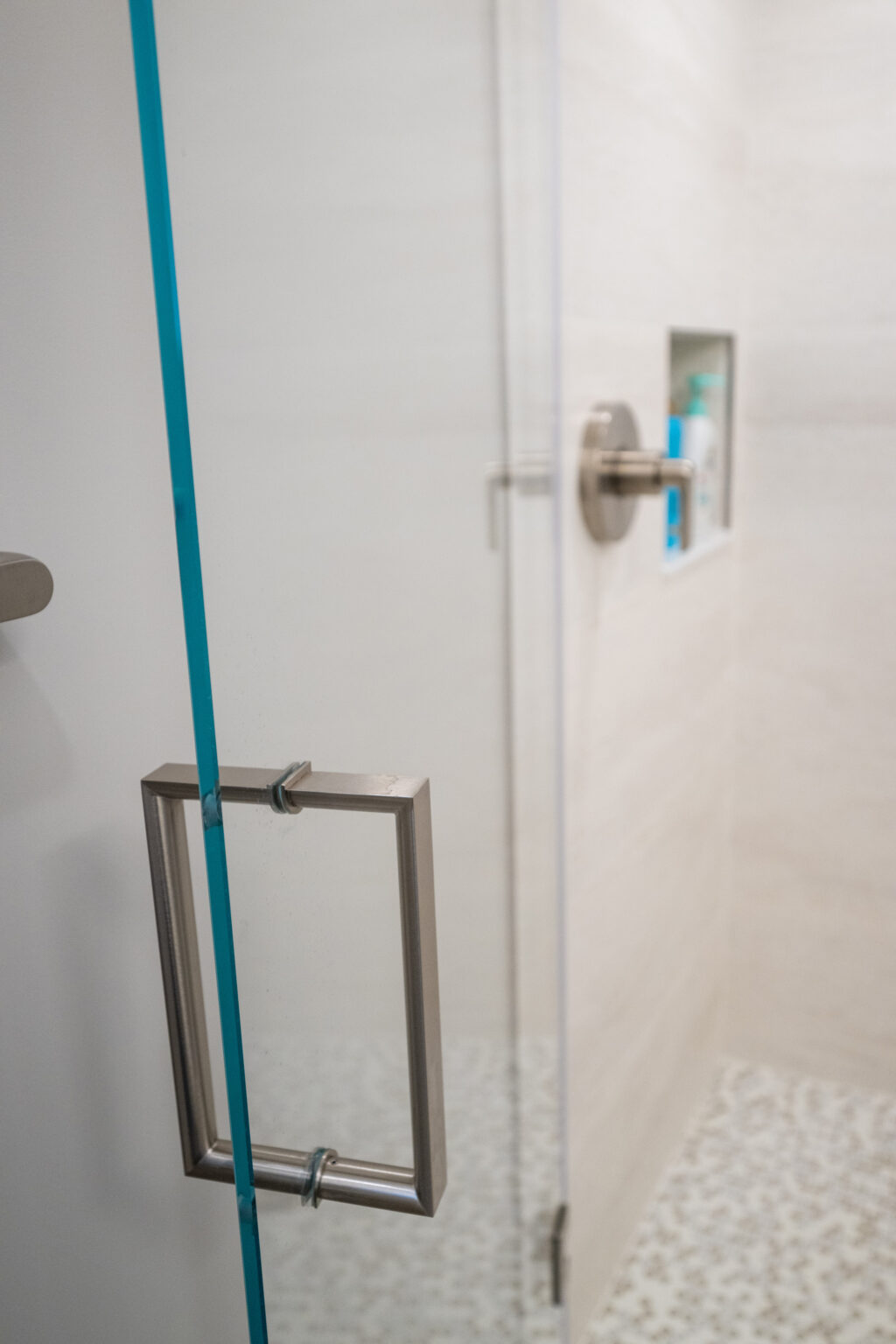 A close-up of a modern shower with a transparent glass door, metal handle, wall-mounted showerhead, and built-in shelf holding toiletries. The shower floor has a pebble-like texture.