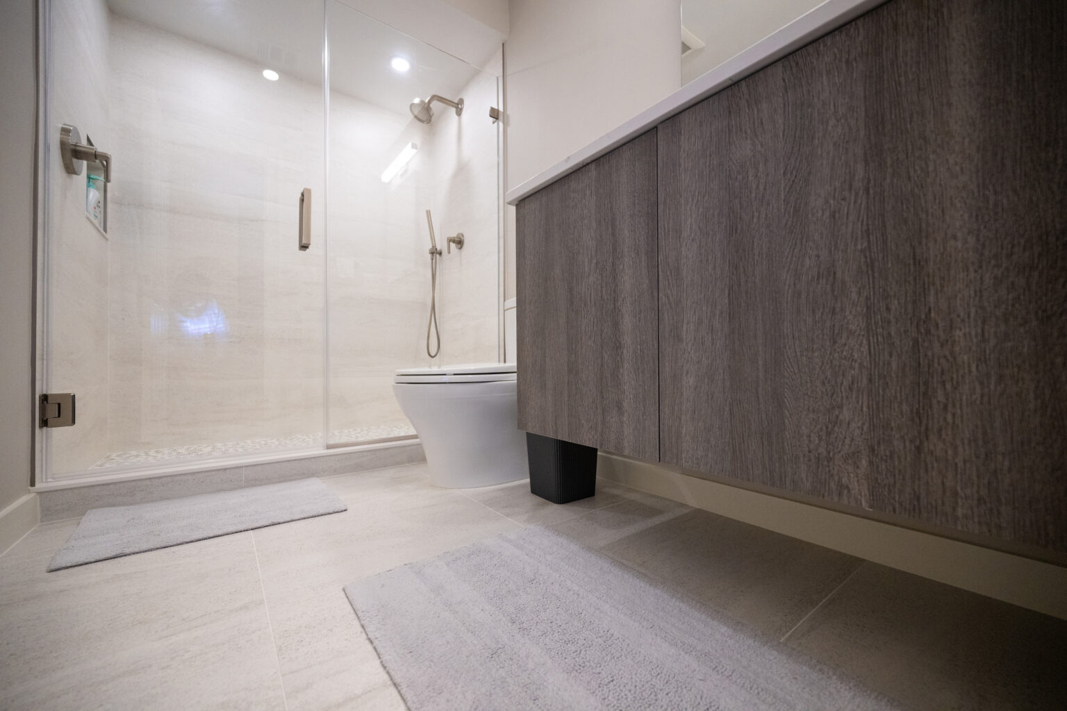 A modern bathroom featuring a glass-enclosed shower, a toilet, a wooden cabinet, and two floor mats. The room has a light color scheme and a minimalist design.