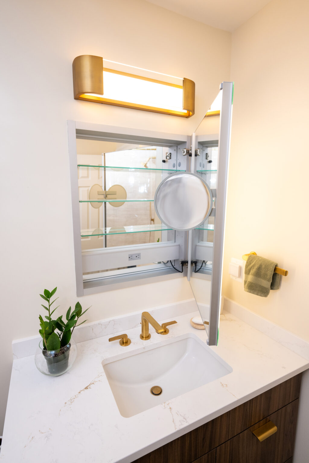 Bathroom countertop with a white sink, brass faucet, mirror with side lights, small round mirror, plant, and a green towel on a rack.