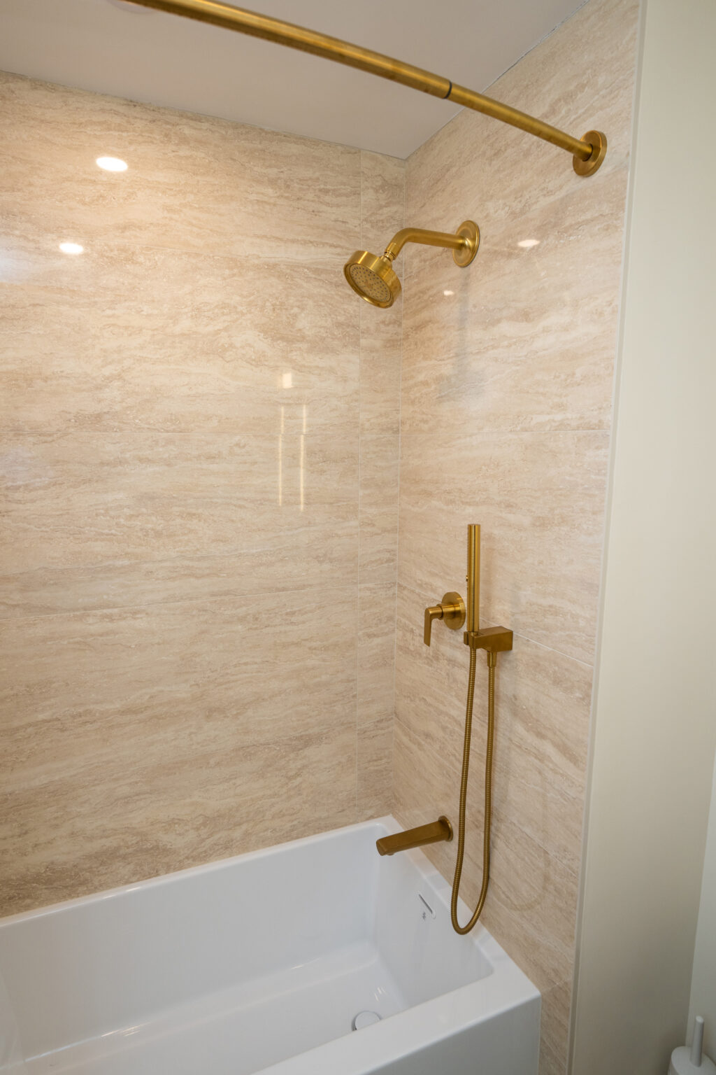 A modern bathroom with a white bathtub and gold fixtures, including a wall-mounted showerhead and faucet, and beige marble walls.