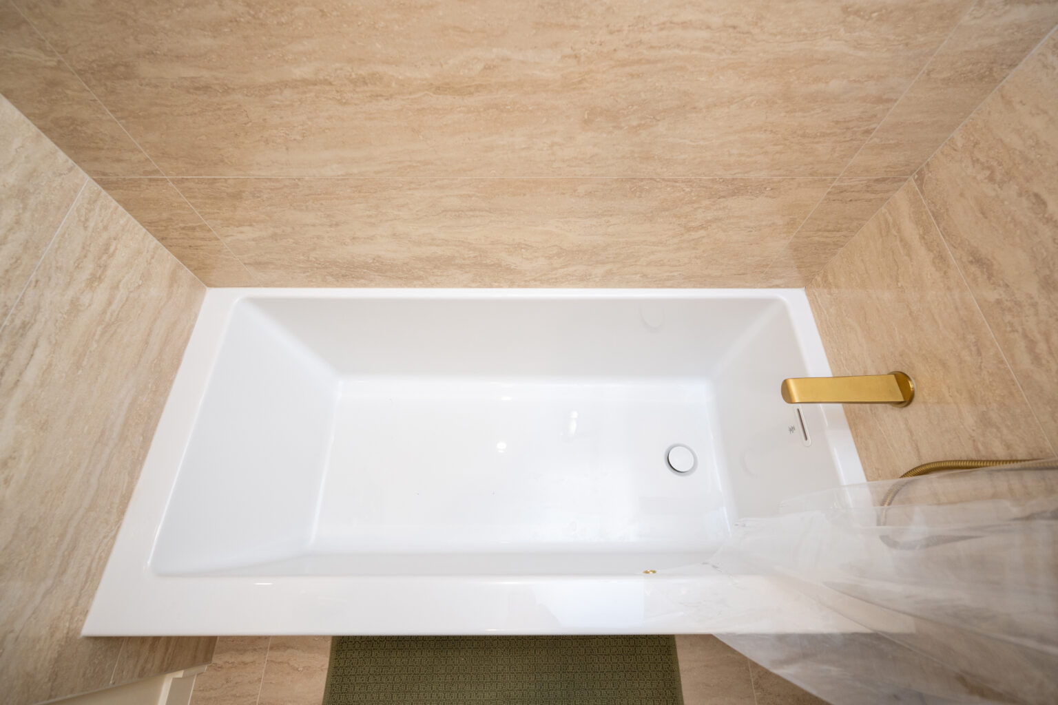 Top-down view of a white rectangular bathtub with a gold faucet fixture, set against light beige tiled walls. A partially visible green bath mat is on the floor next to the tub.