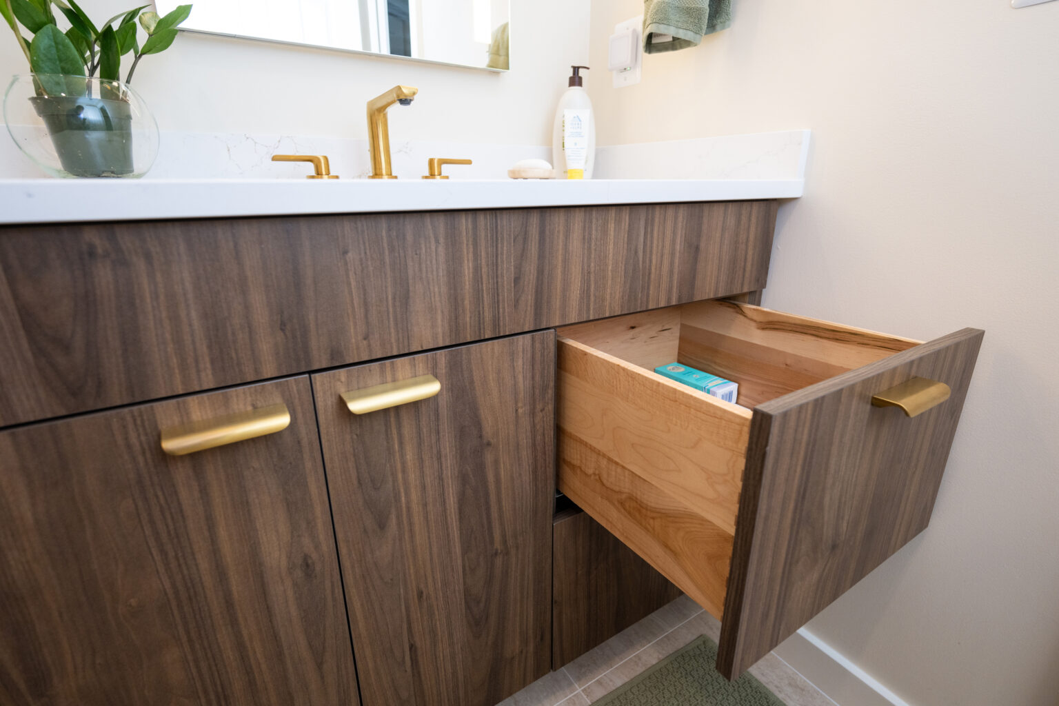 A bathroom vanity with a wood finish is shown. One drawer is open, revealing items inside. The countertop has a gold faucet, a small potted plant, and various toiletries.