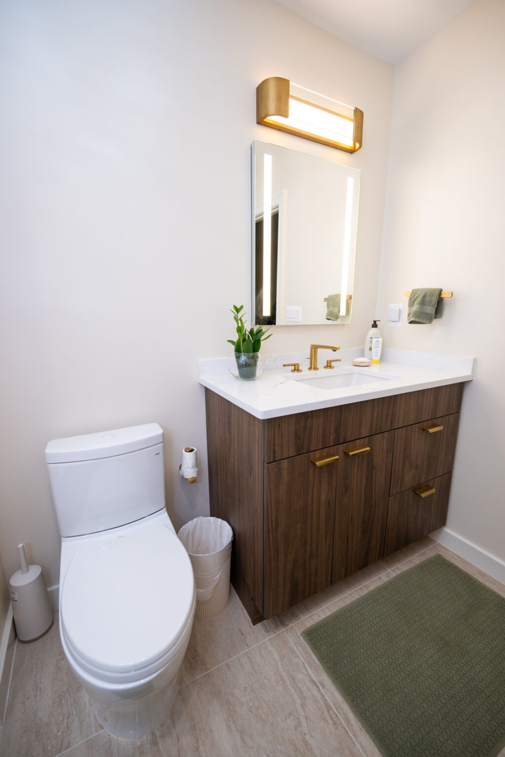 A modern bathroom featuring a toilet, a wooden vanity with a white sink, a mirror with a light fixture, and a green mat on the floor. A potted plant and soap dispenser are on the counter.