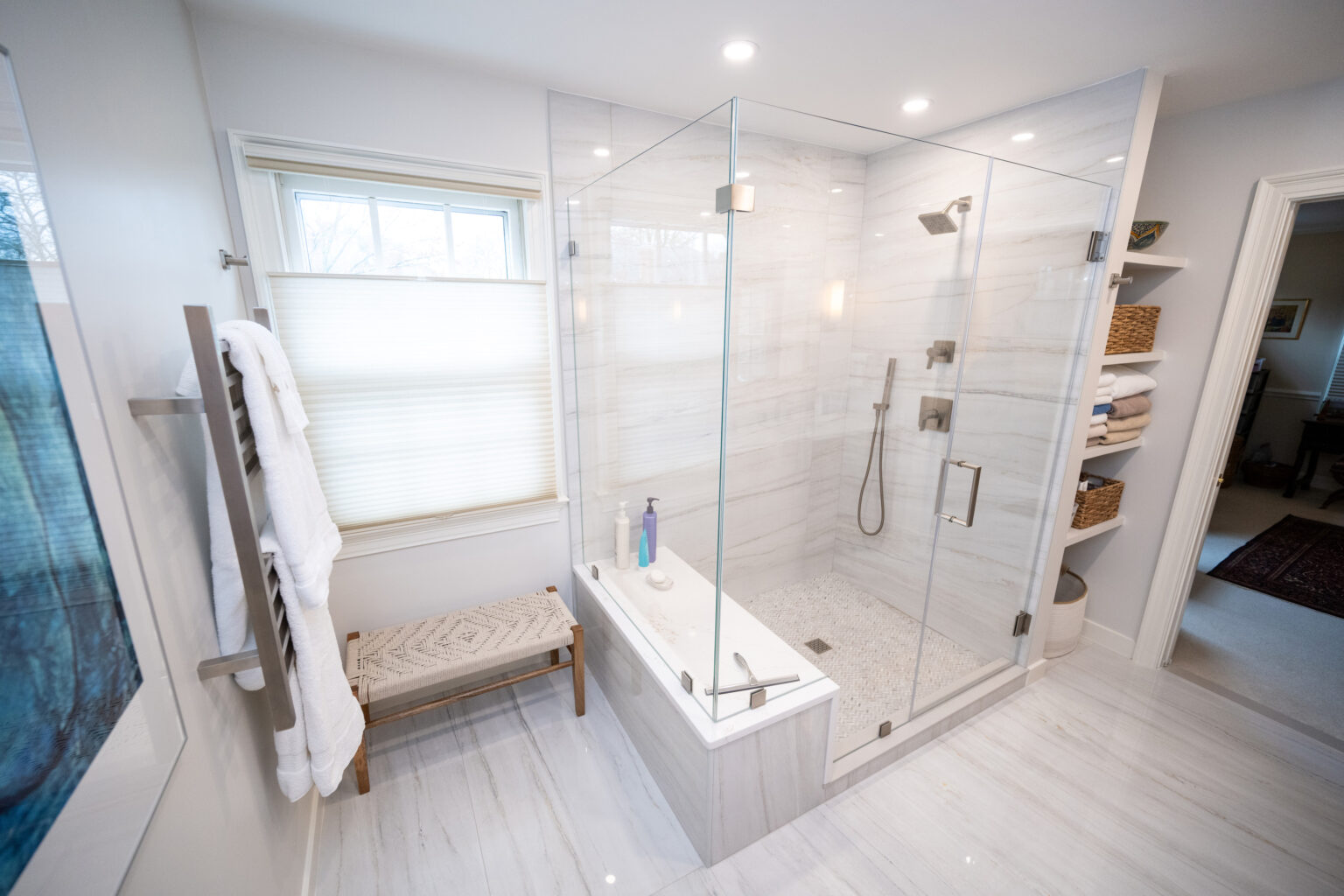 Modern bathroom with a glass-enclosed shower next to a bathtub, a towel rack with folded towels, and a bench. Light tiled floors and walls, and shelves with wicker baskets are visible.