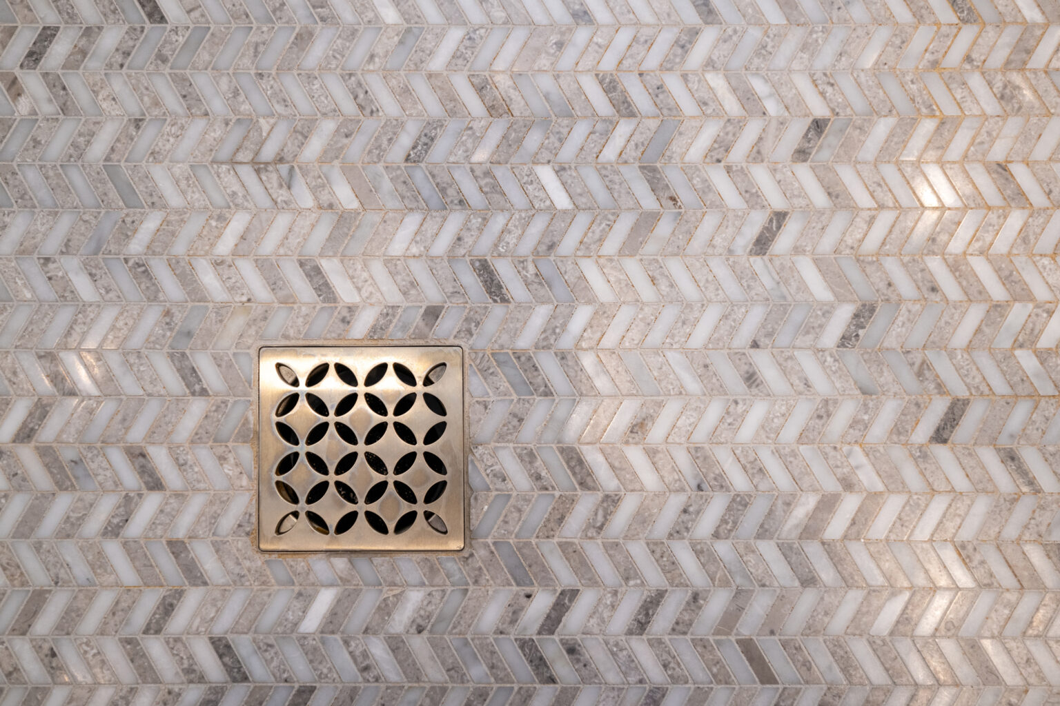 Close-up of a square metallic drain cover with a floral cutout pattern on a herringbone marble tile floor.
