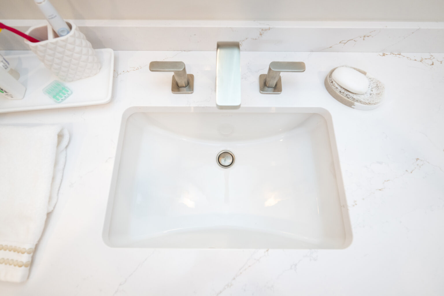 A bathroom sink with a modern faucet, a toothbrush holder, soap, and a neatly folded towel on the white countertop.