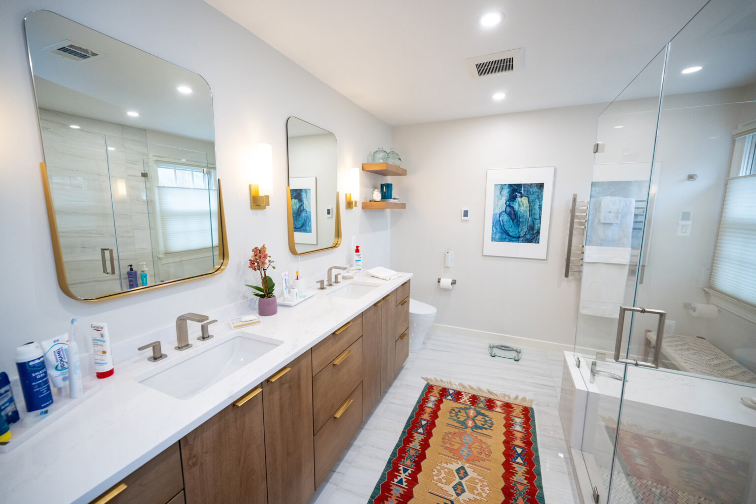 Modern bathroom with double sinks, large mirrors, wooden cabinets, glass shower, and a colorful rug. Decor includes potted plants, shelves, and framed artwork on the wall. Well-lit with ceiling lights.