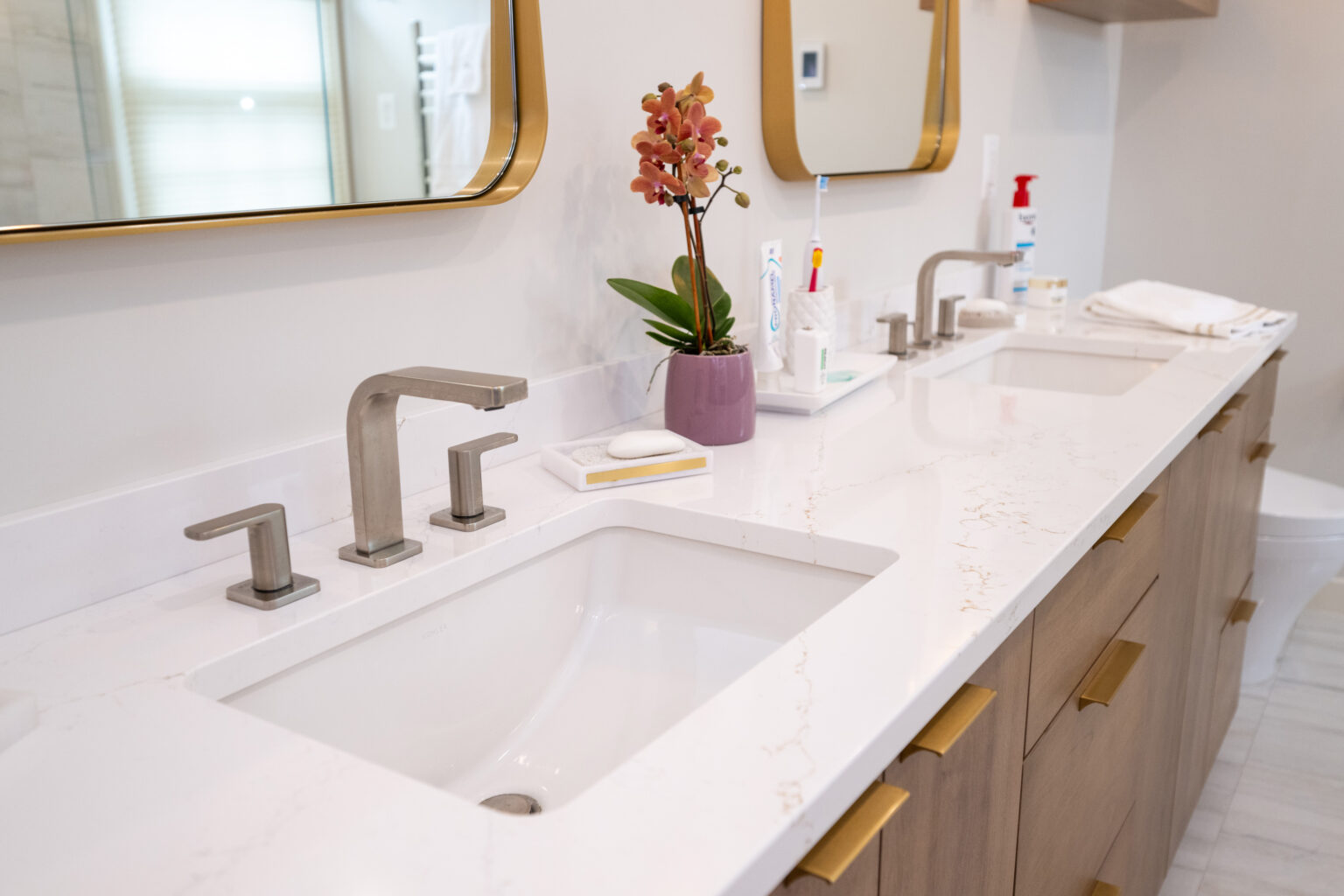 A modern bathroom with a double sink vanity, orchid plant, soap, toothbrushes, and a hand sanitizer bottle on the countertop. Large mirrors and brass fixtures are mounted above the sinks.