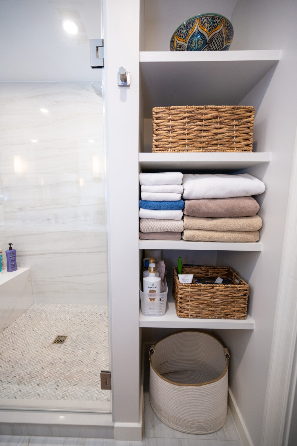 Bathroom shelving unit with wicker baskets, folded towels, and toiletries next to a walk-in shower with a glass door.