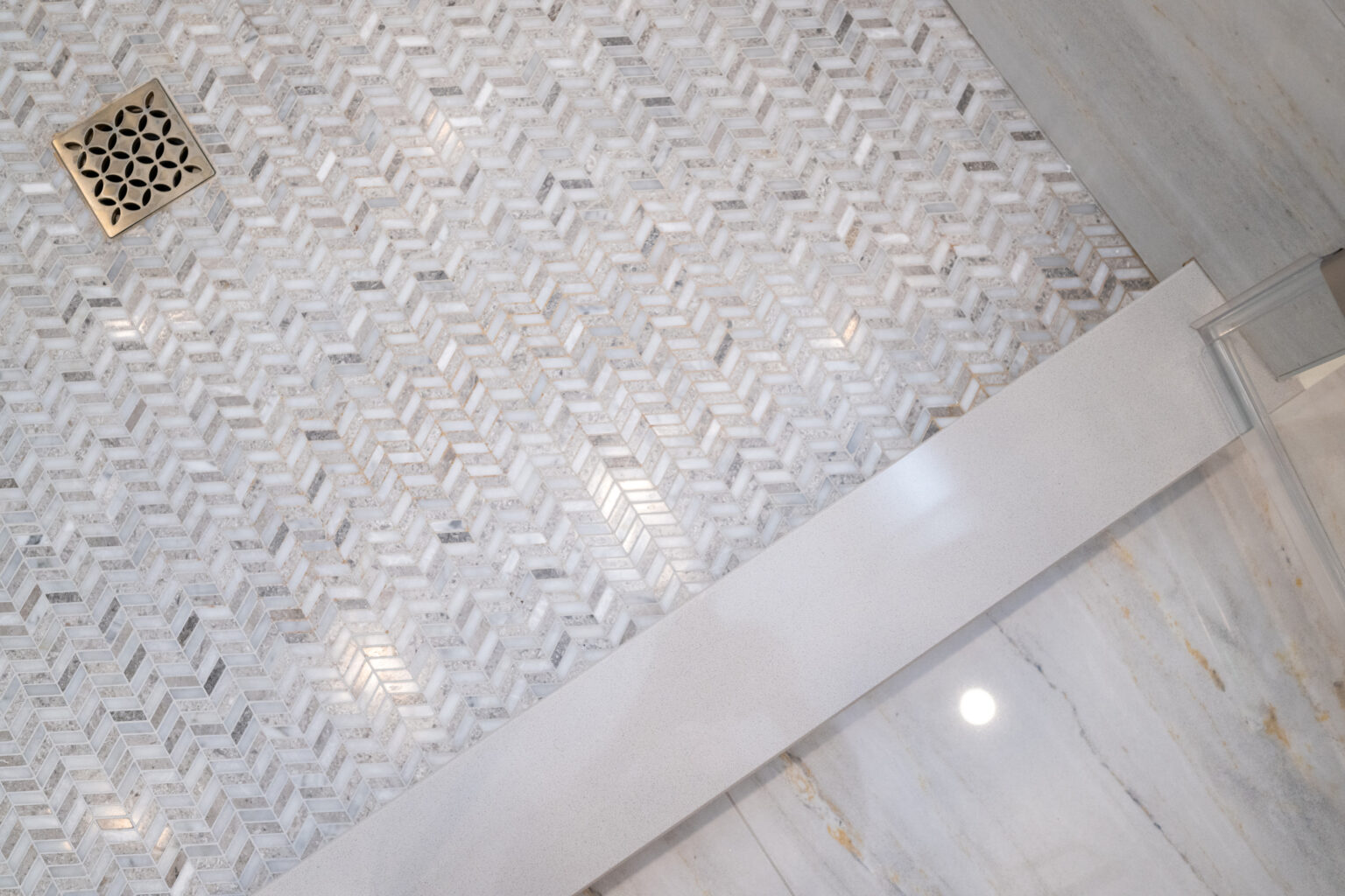 A marble herringbone mosaic tile floor with a metal drain cover in the upper left corner. The flooring features white and gray tones.