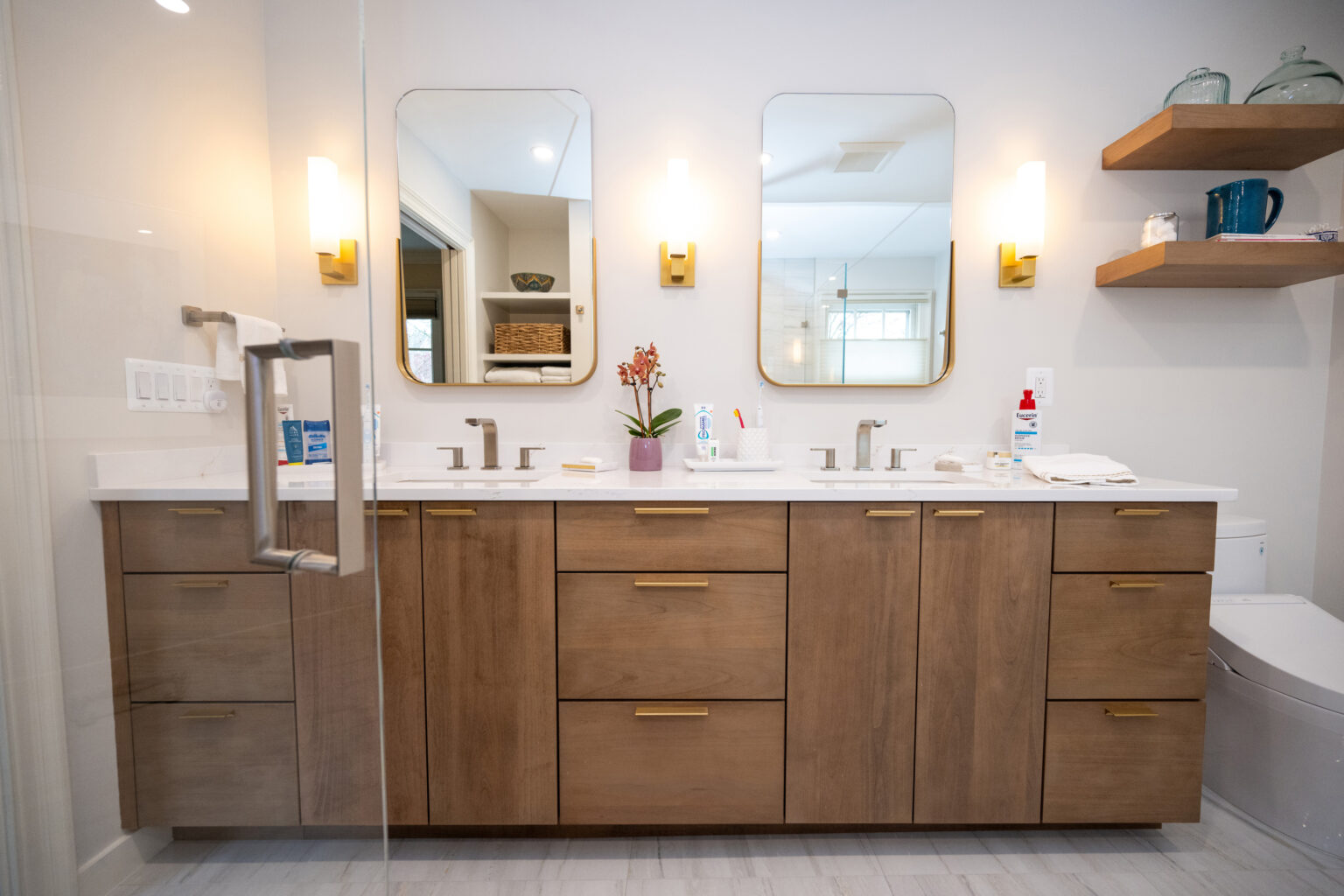 A modern bathroom features a double sink vanity with wooden cabinets and brass handles. Two mirrors are mounted above, flanked by wall sconces. A toilet and shelves with decor items are visible.